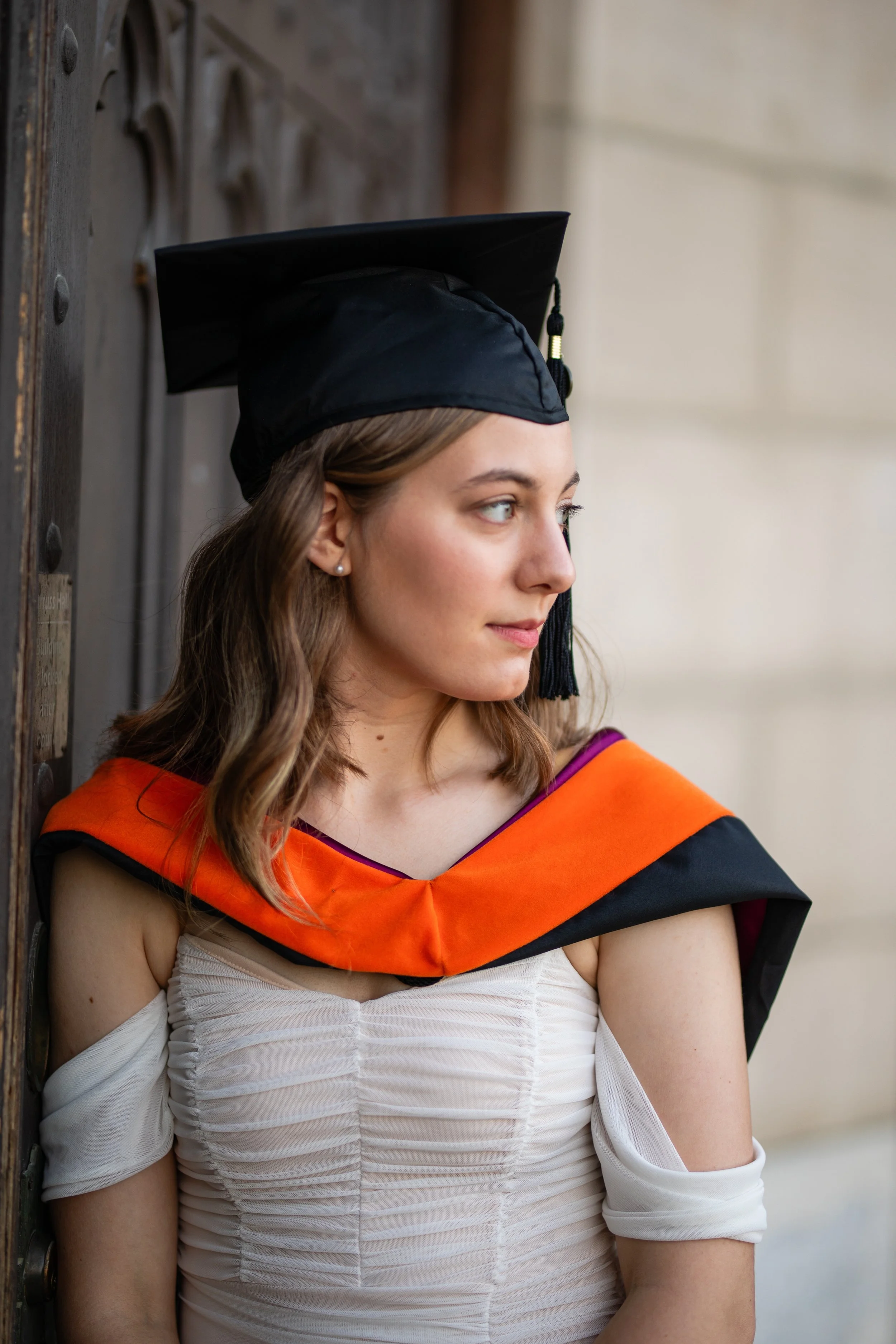 A young woman in a white dress and black graduation cap with an orange and black academic hood, standing outdoors and looking to the side.