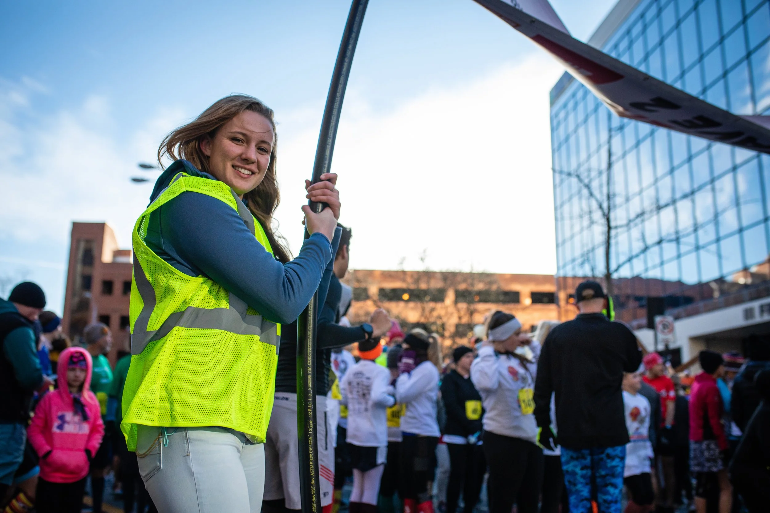 A young woman smiling at camera, wearing a yellow reflective safety vest, holding a flag at an outdoor event with people gathered in the background.
