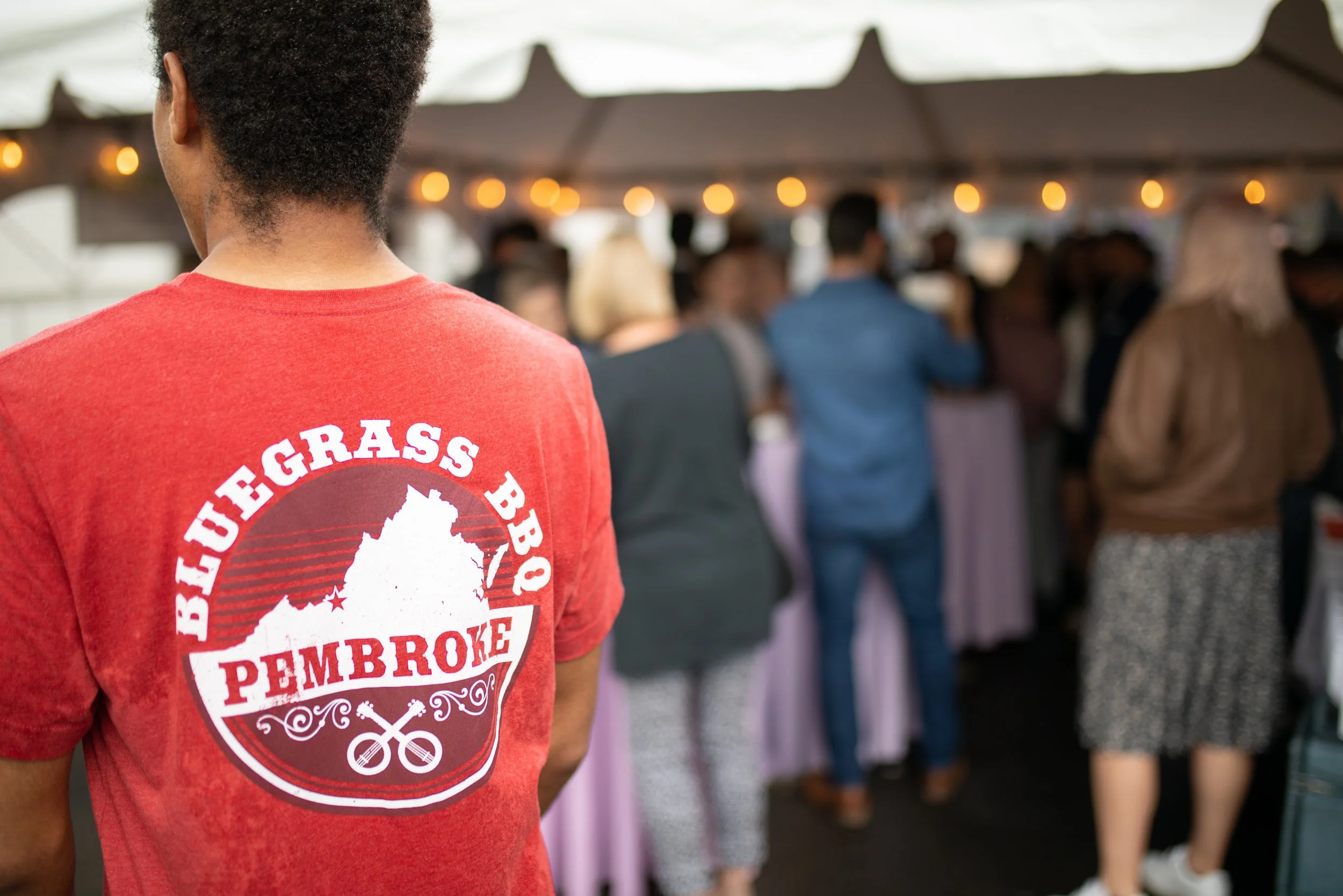 Person wearing a red T-shirt with the logo for Bluegrass BBQ Pembroke, standing in front of a crowd at a barbecue event under a large tent with string lights.