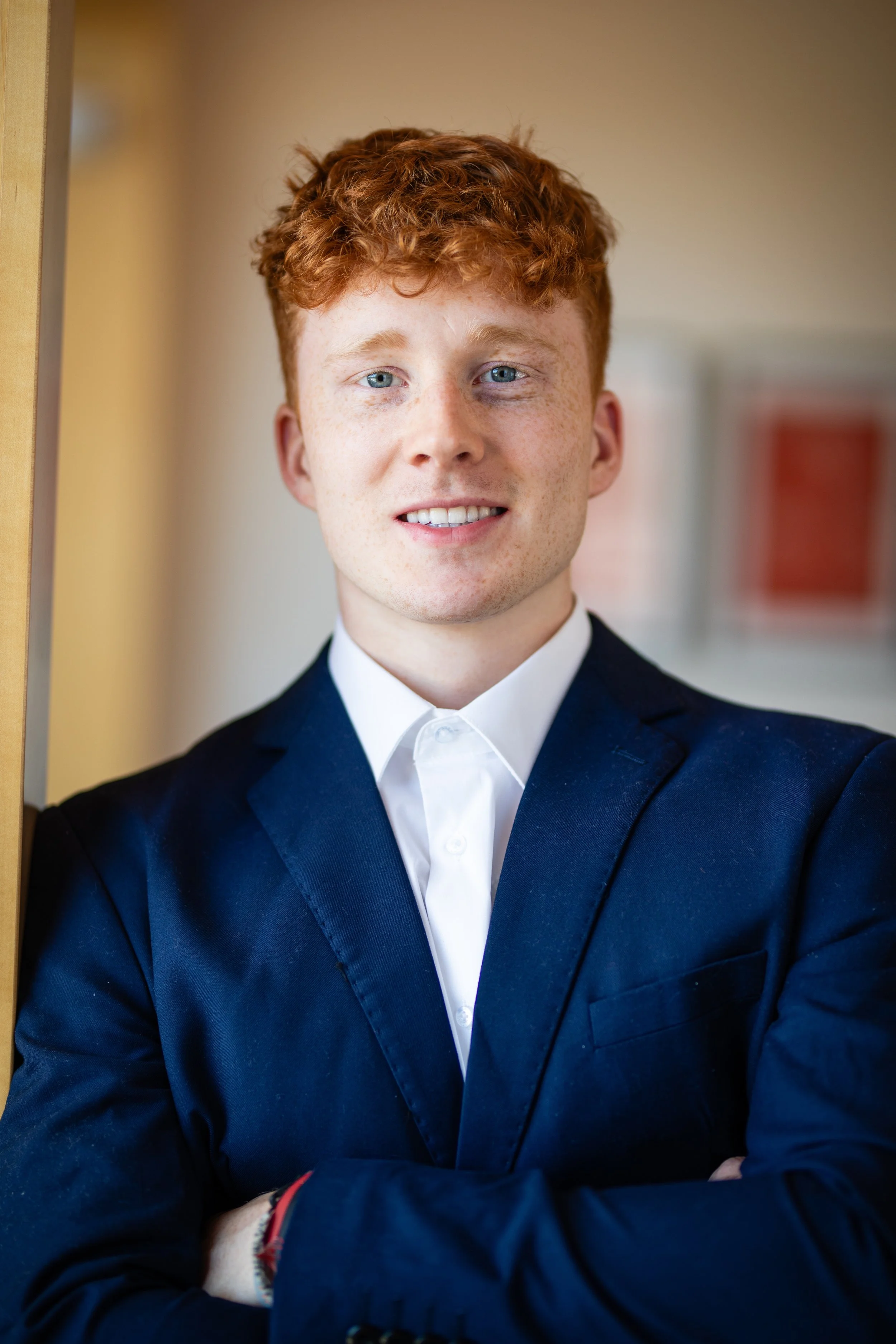 Young man with red hair, blue eyes, wearing a white shirt and navy blazer, standing with arms crossed.