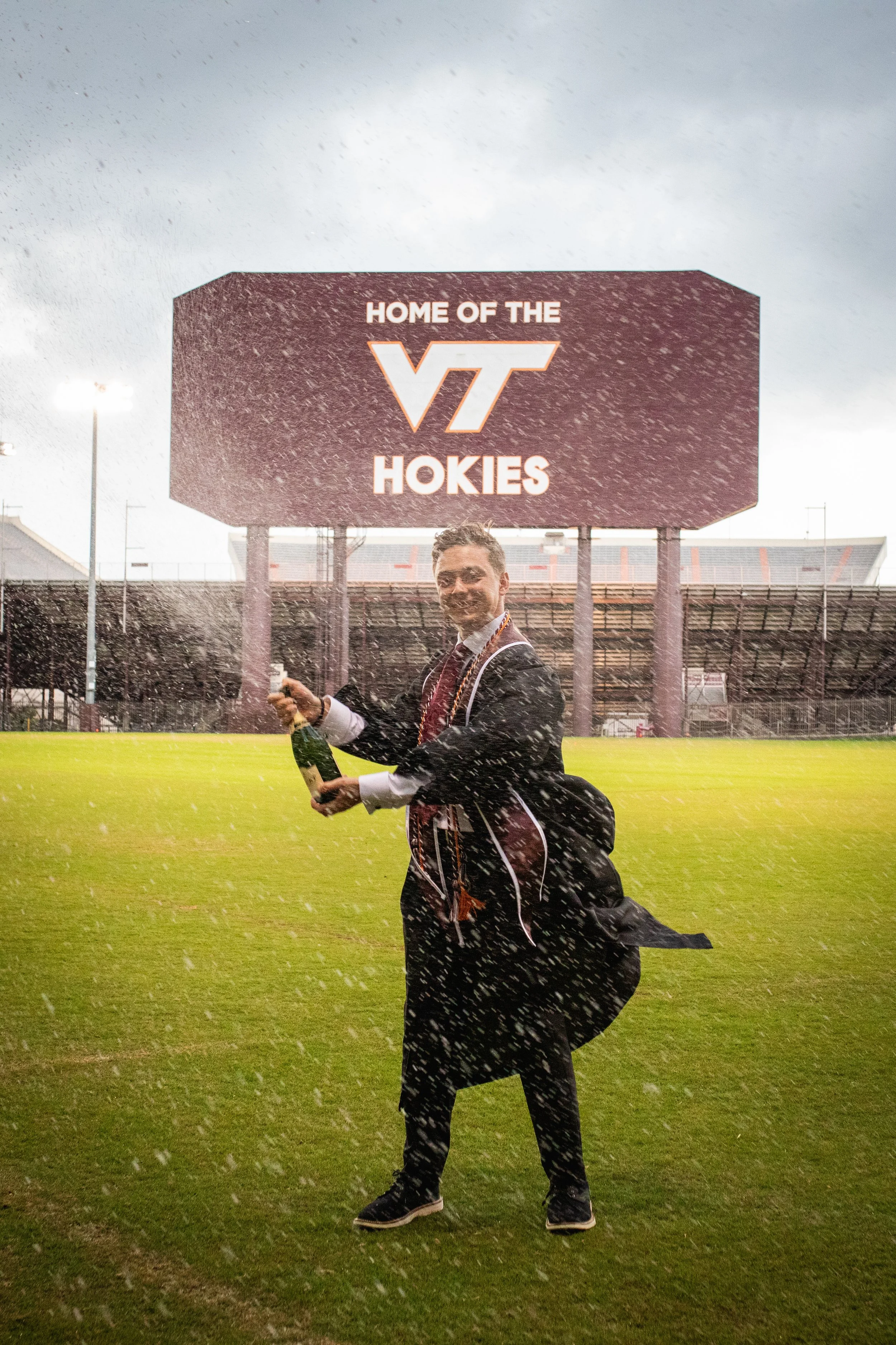 A person in academic regalia celebrating on a football field holding a bottle of champagne while spraying champagne in the air. Behind him is a sign that reads 'Home of the VT Hokies' and a large stadium.