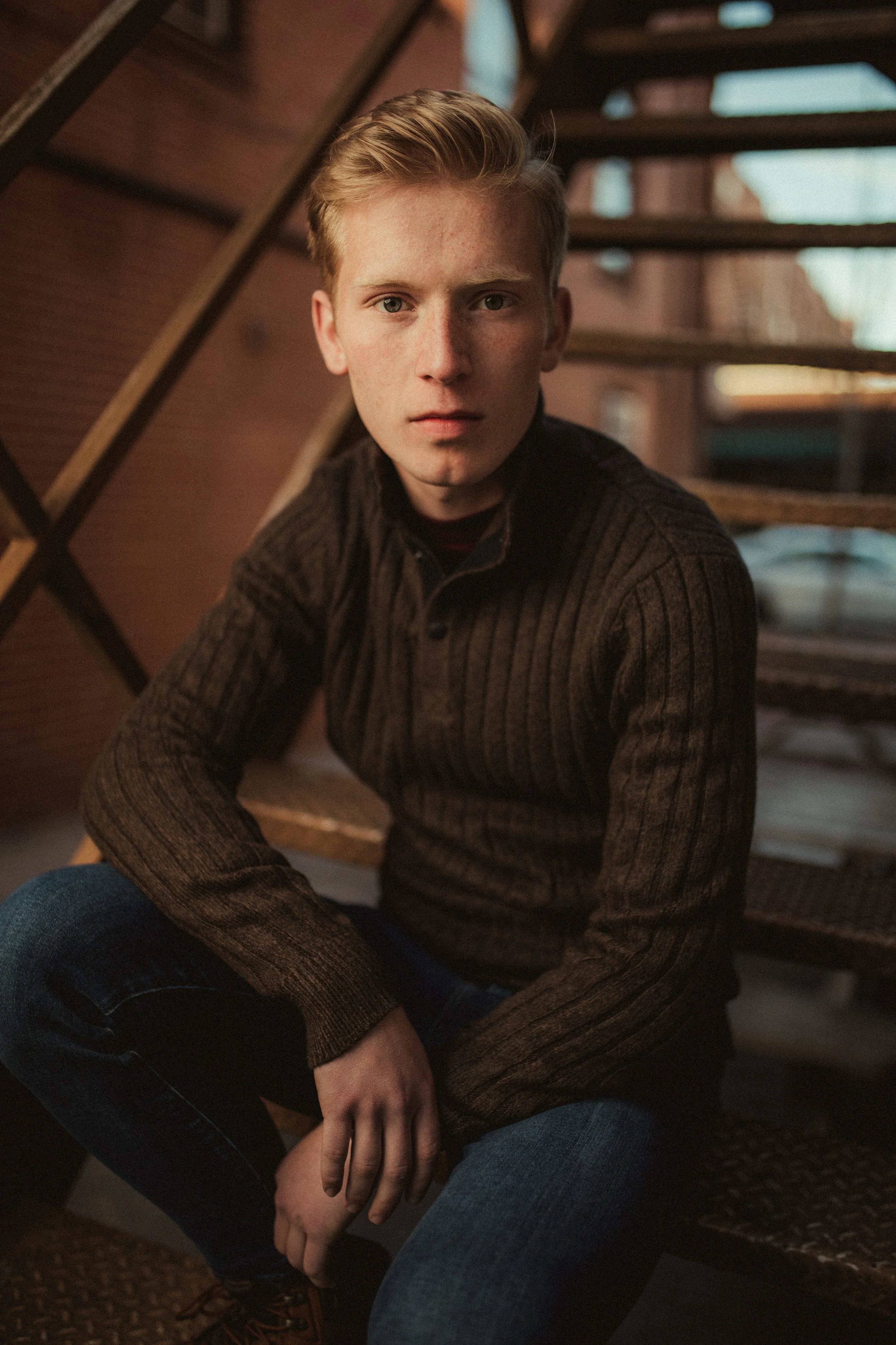 A young man with blond hair, wearing a brown sweater and jeans, sitting on metal stairs inside a building with wooden walls and large windows.