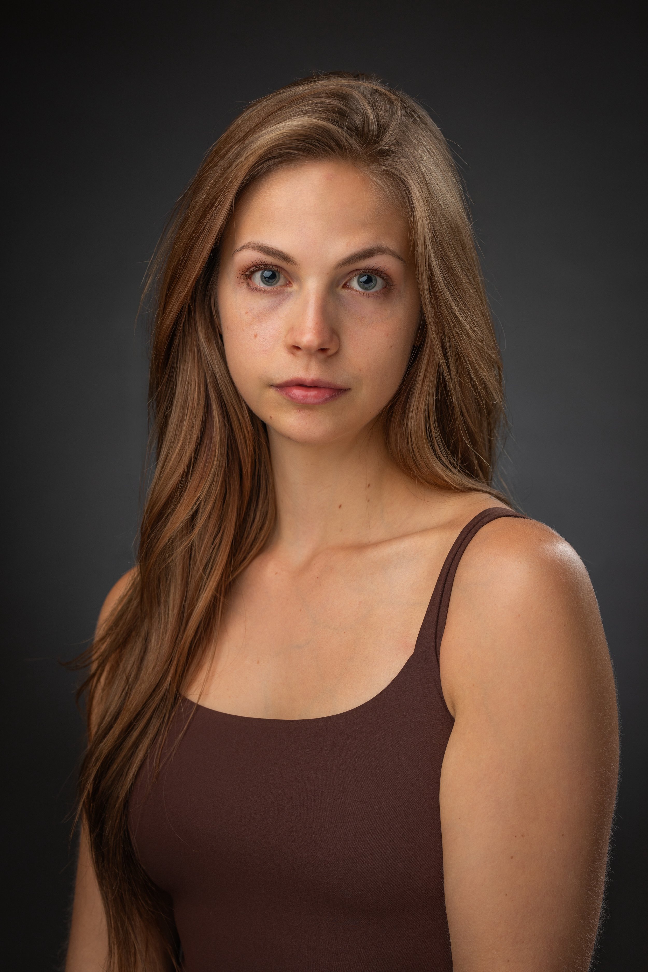 Portrait of a young woman with long, wavy brown hair wearing a brown tank top, against a dark background.