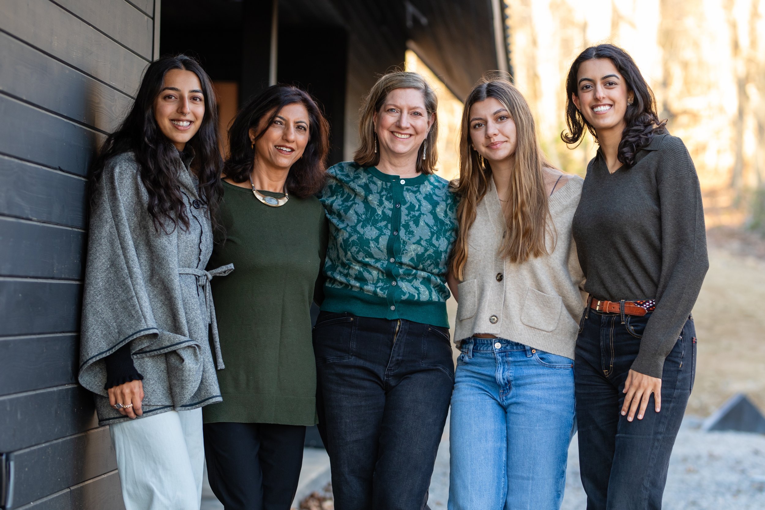 Group of five women standing close together outdoors, smiling, dressed casually. One woman leans against a dark wooden wall, and the background shows trees and golden light.