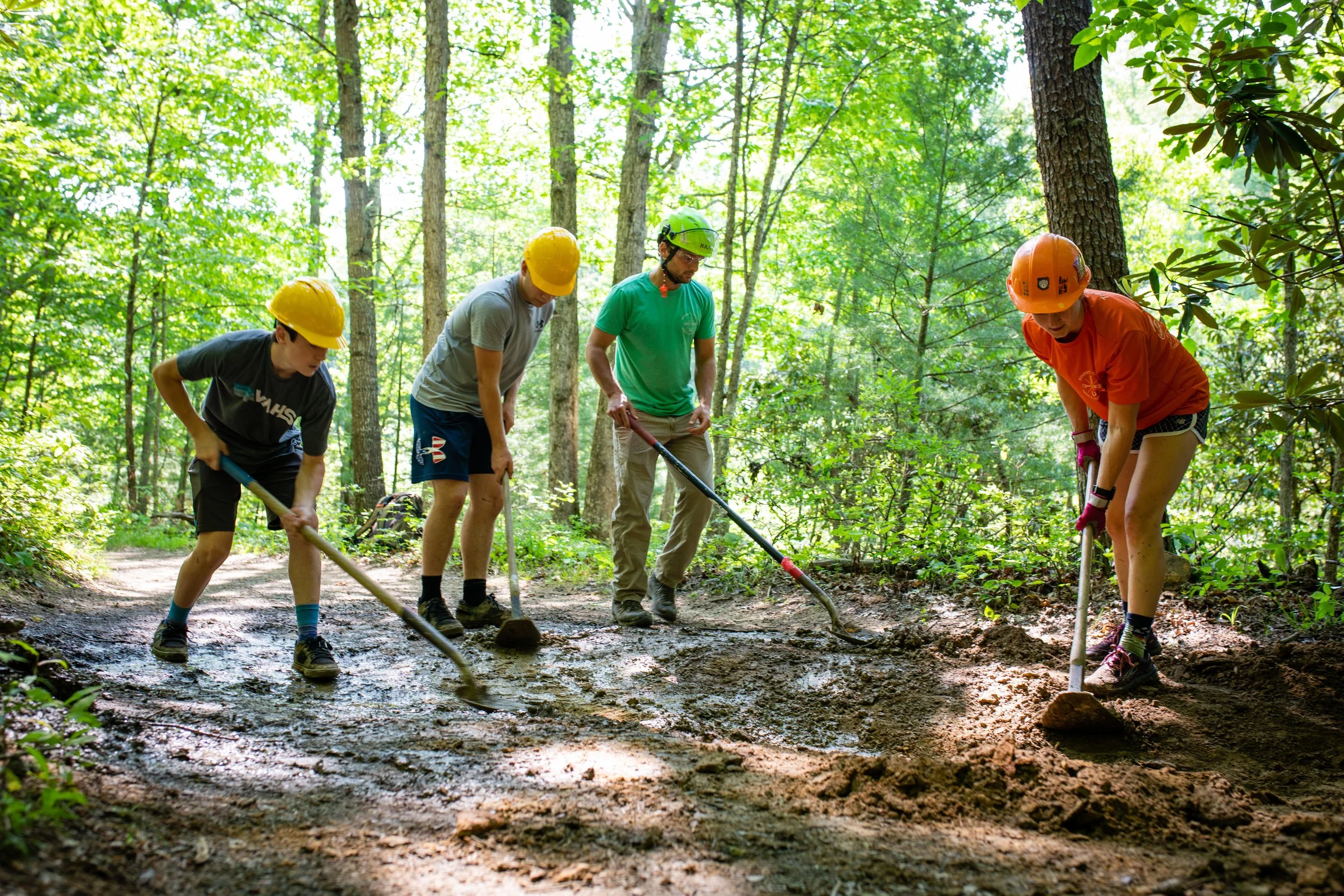 Group of four people wearing helmets and working with shovels on a trail in a lush green forest.