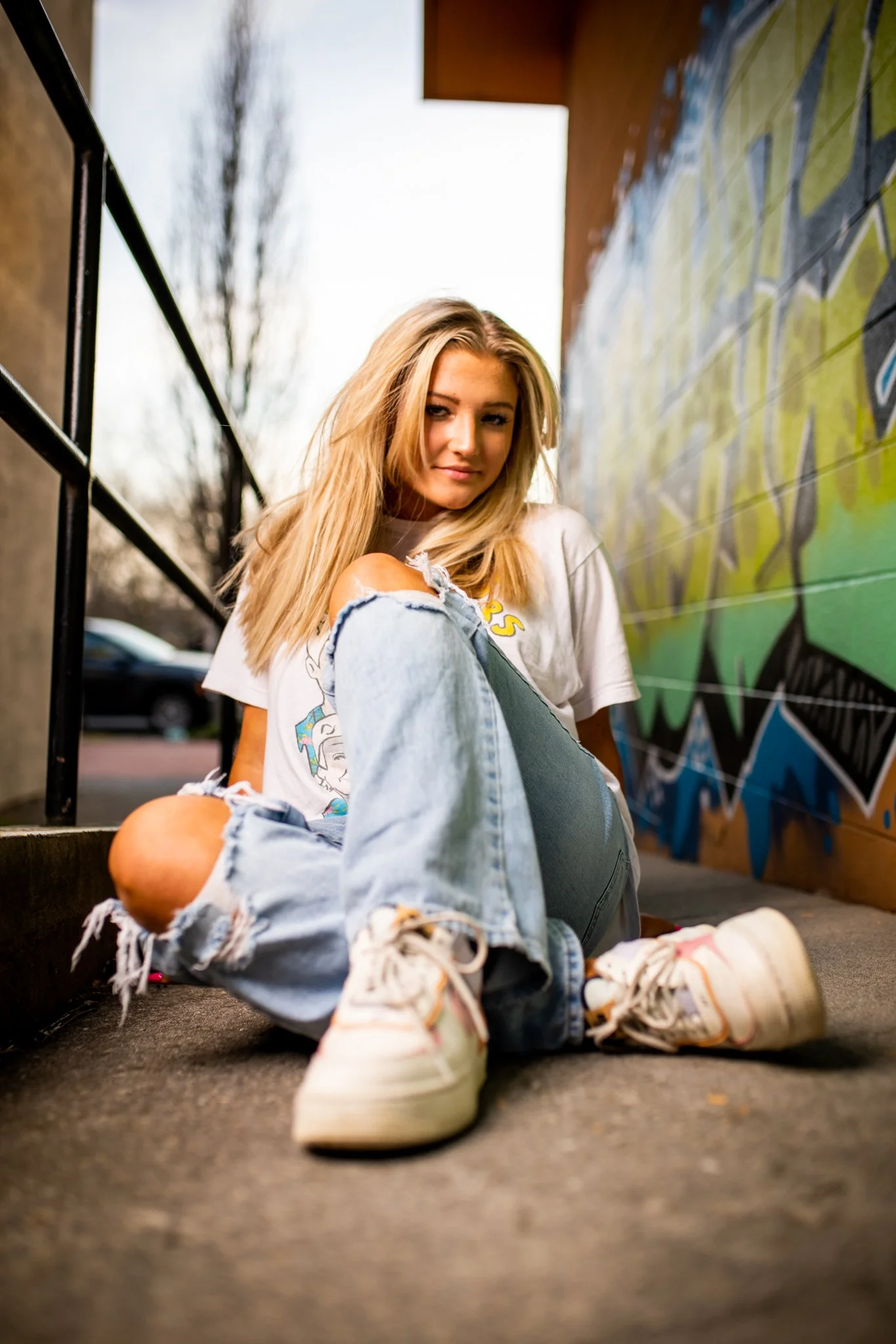 A young woman with blonde hair wearing a white graphic T-shirt and ripped jeans sits on the ground near a colorful graffiti wall, outdoors during daytime.