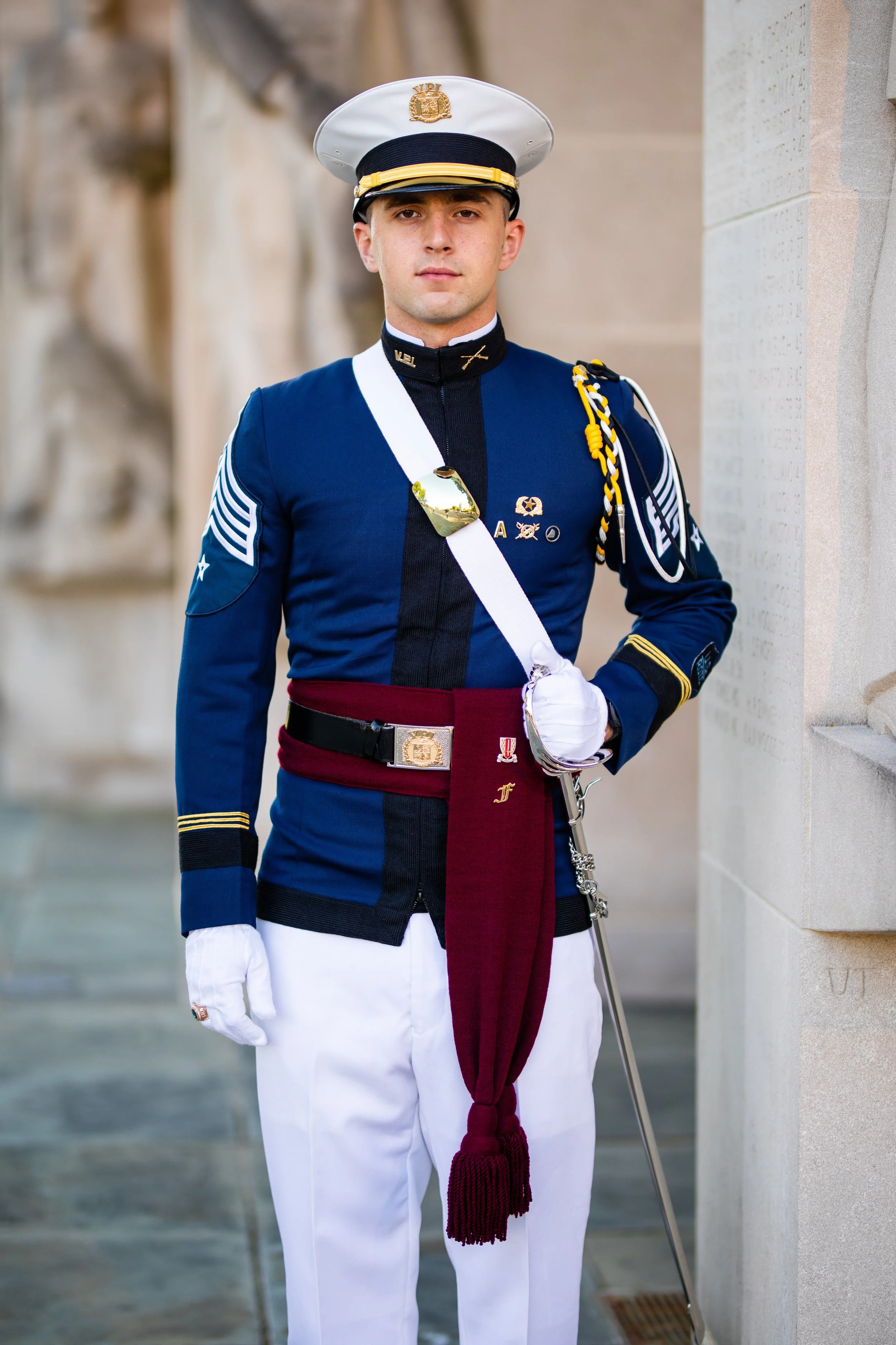 A uniformed member of the U.S. military standing outdoors, wearing a blue dress uniform with medals, a white hat with a gold insignia, white gloves, and holding a ceremonial sword.