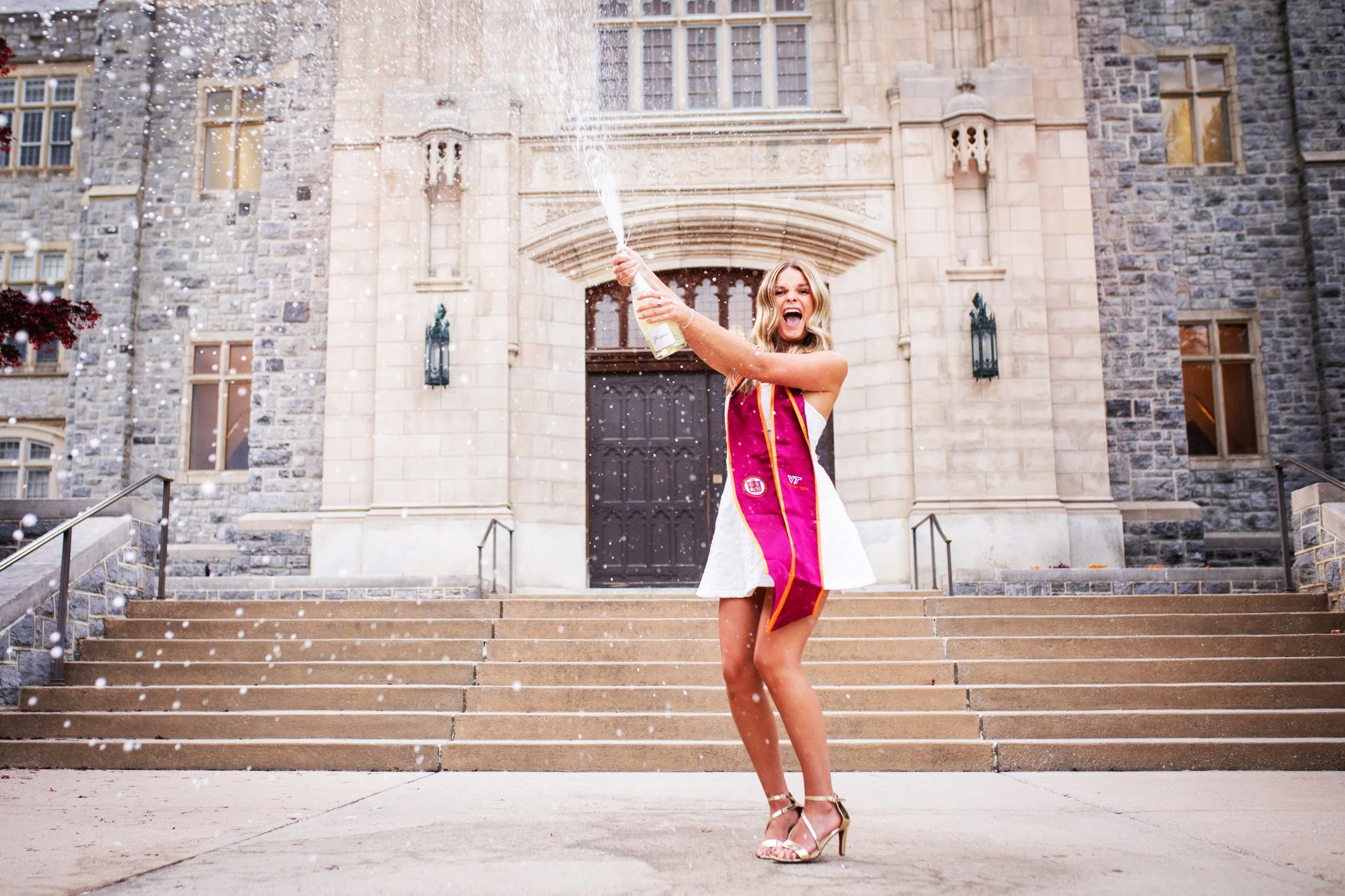 A woman in a white dress and graduation stole celebrating on campus with a champagne bottle in hand, spraying it in the air while standing on the steps of a large stone building.