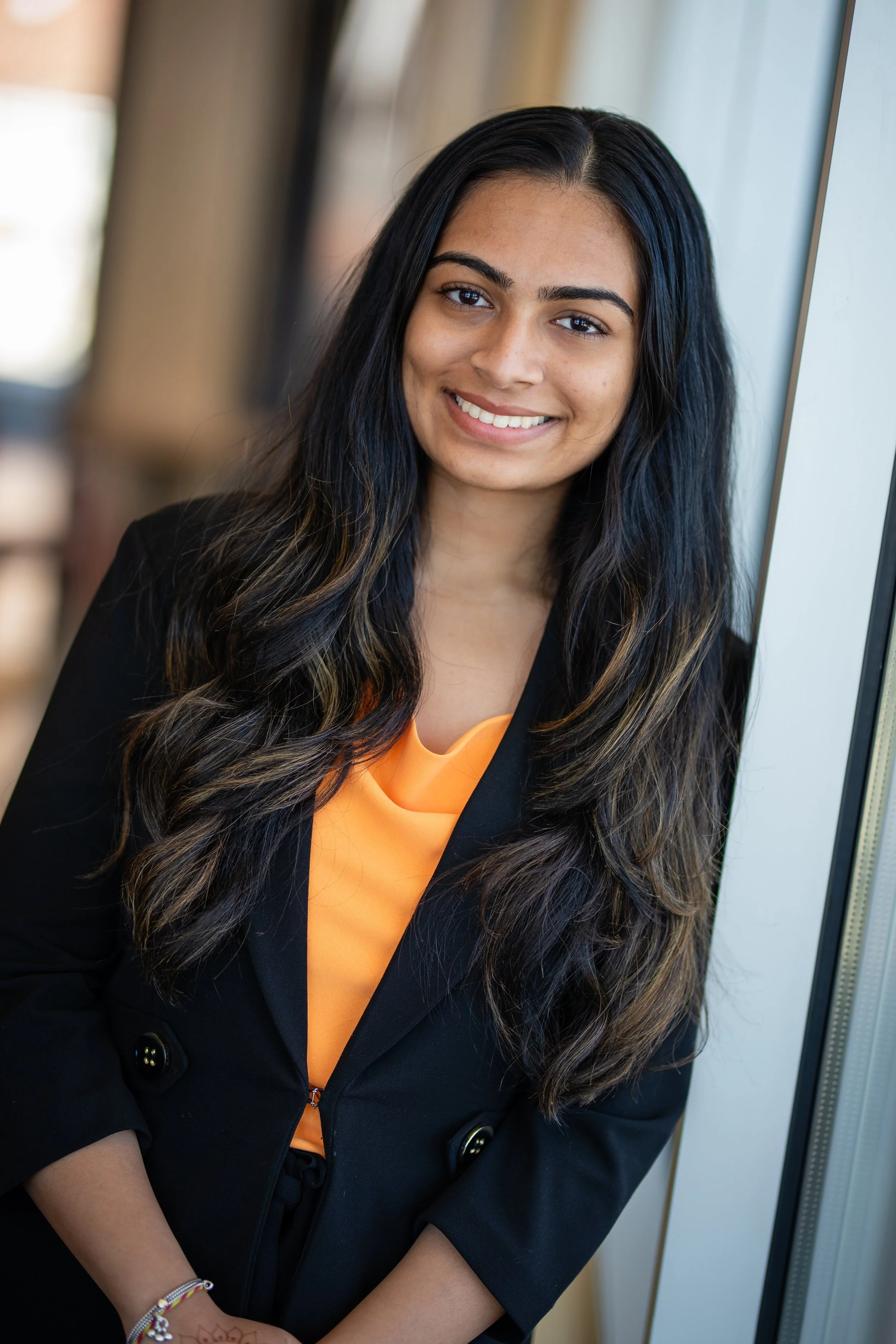 A young woman with long wavy black hair, wearing a black blazer and an orange top, smiling and standing next to a window.
