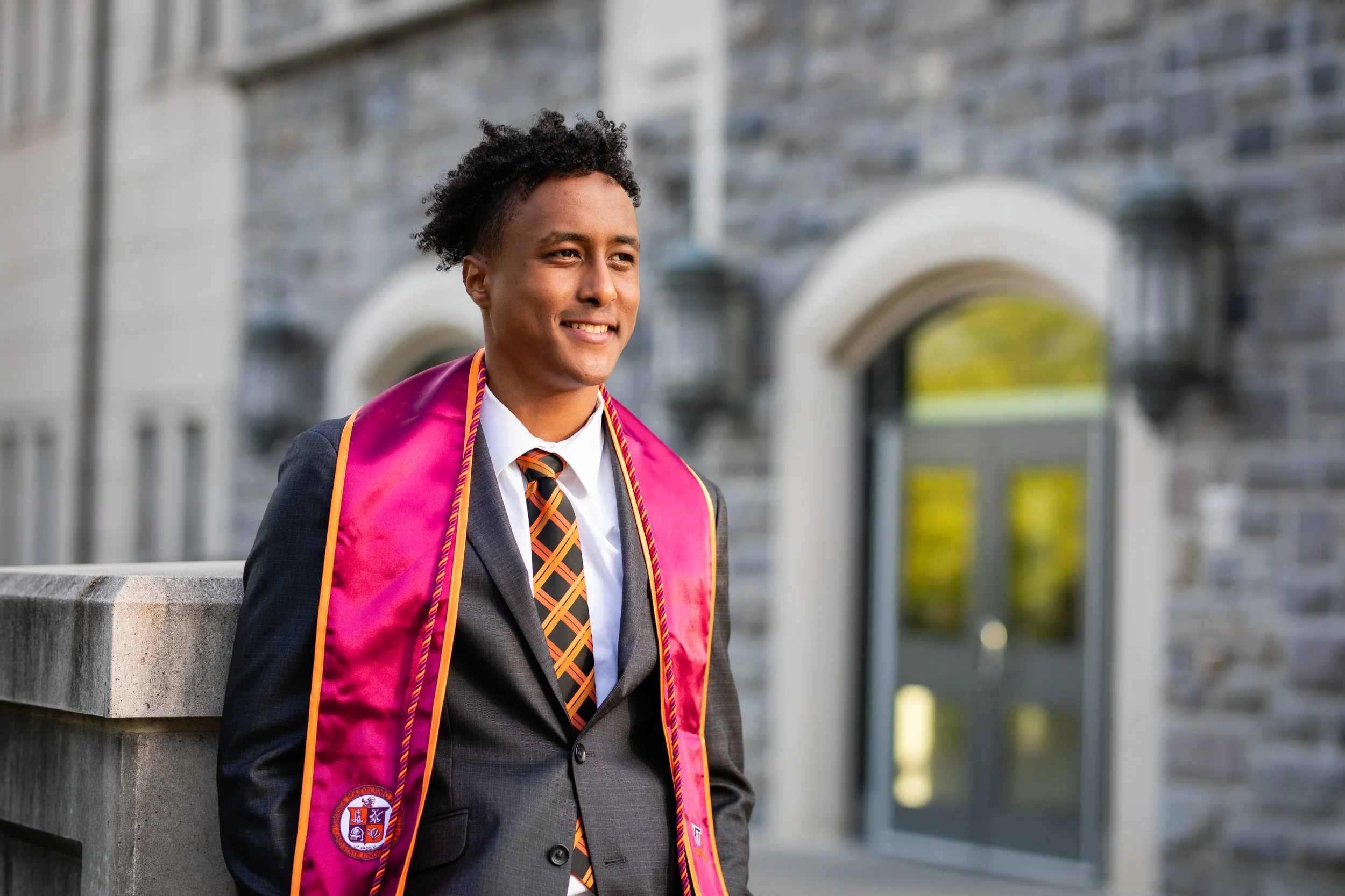 A young man in a graduation gown and a suit with an embroidered pink stole and tie, smiling outdoors near a stone building.