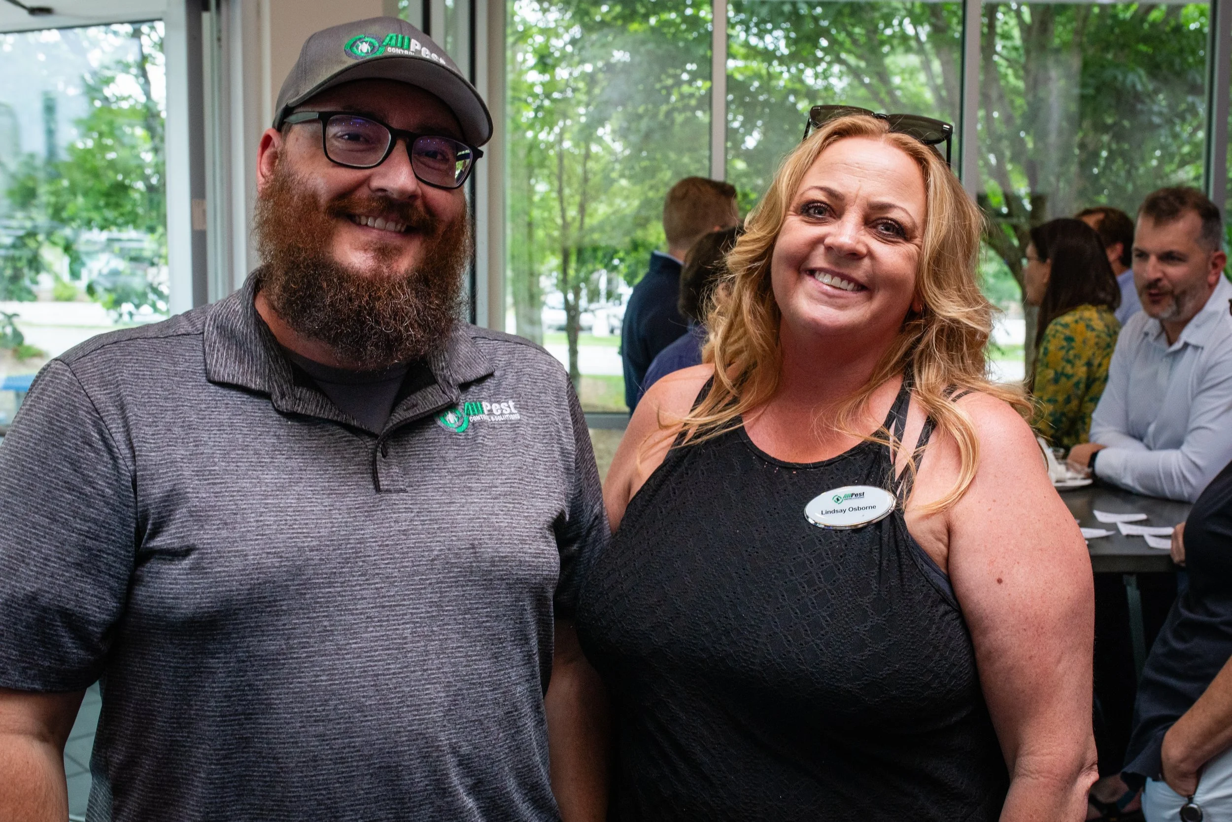 Smiling man with a beard wearing a gray polo shirt and baseball cap standing next to woman with blonde hair in black sleeveless top at a social event.