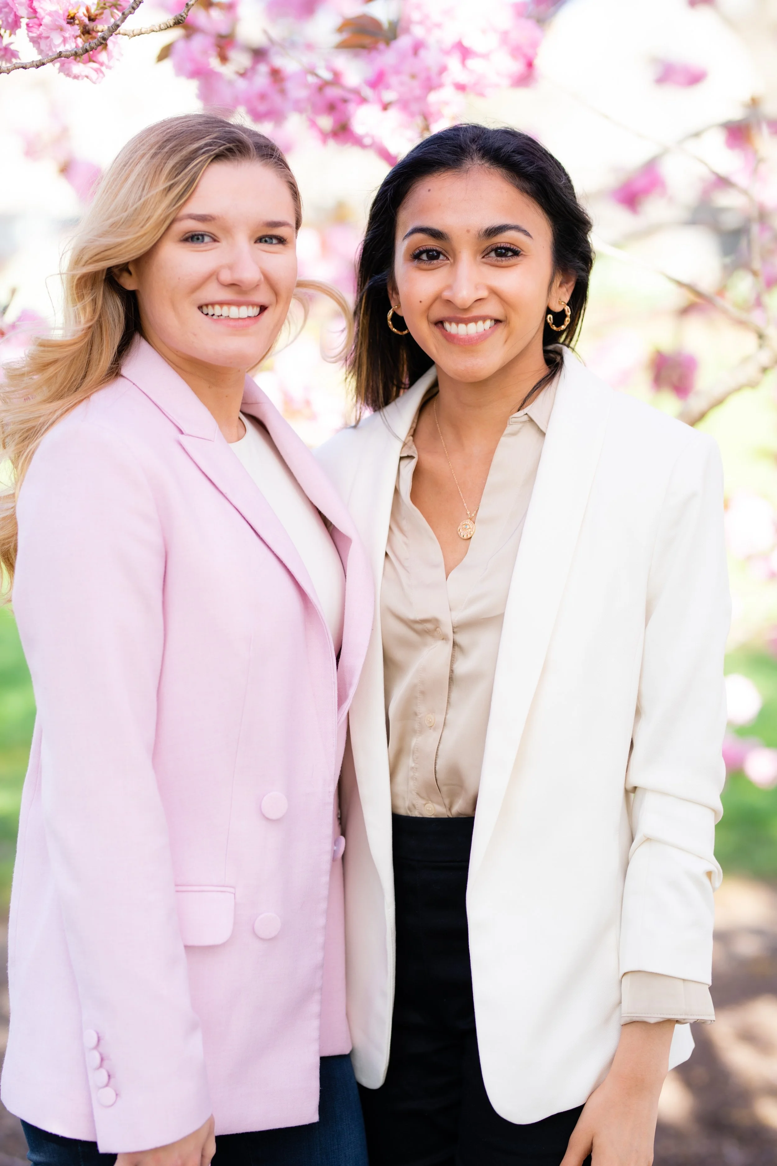 Two women standing close together in front of blooming pink trees, smiling at the camera.