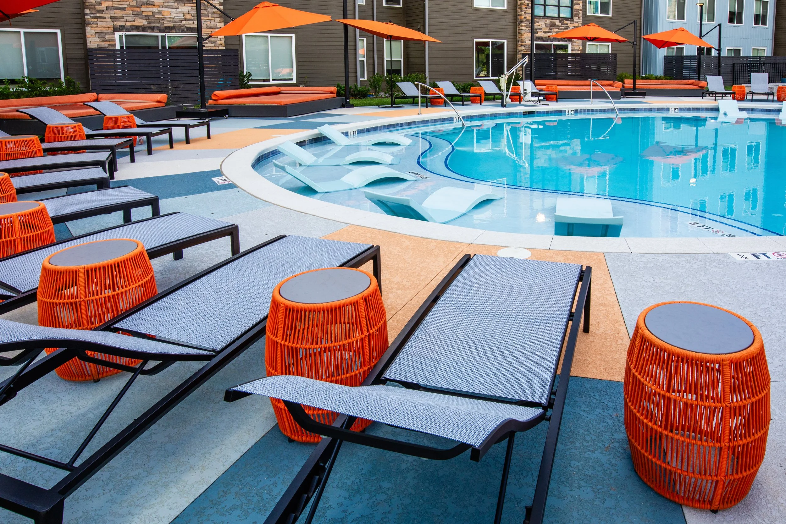 Empty outdoor swimming pool area with lounge chairs, orange side tables, and orange umbrellas in a residential complex.