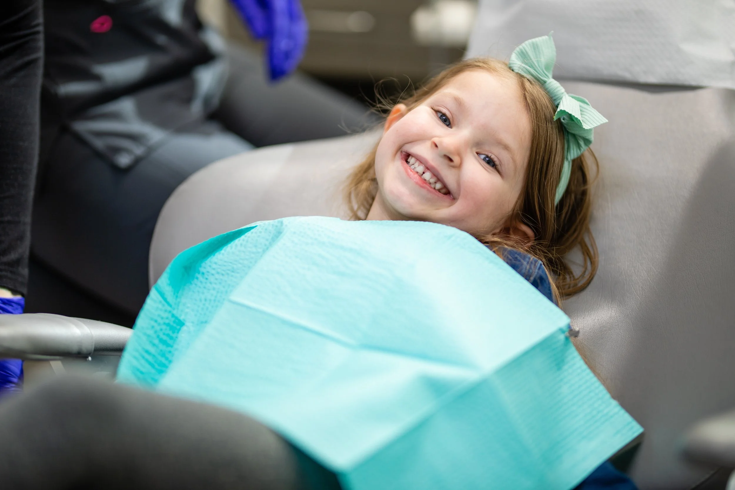 Young girl with long hair and a green bow lying on a dental chair, smiling, with a blue protective bib around her neck.
