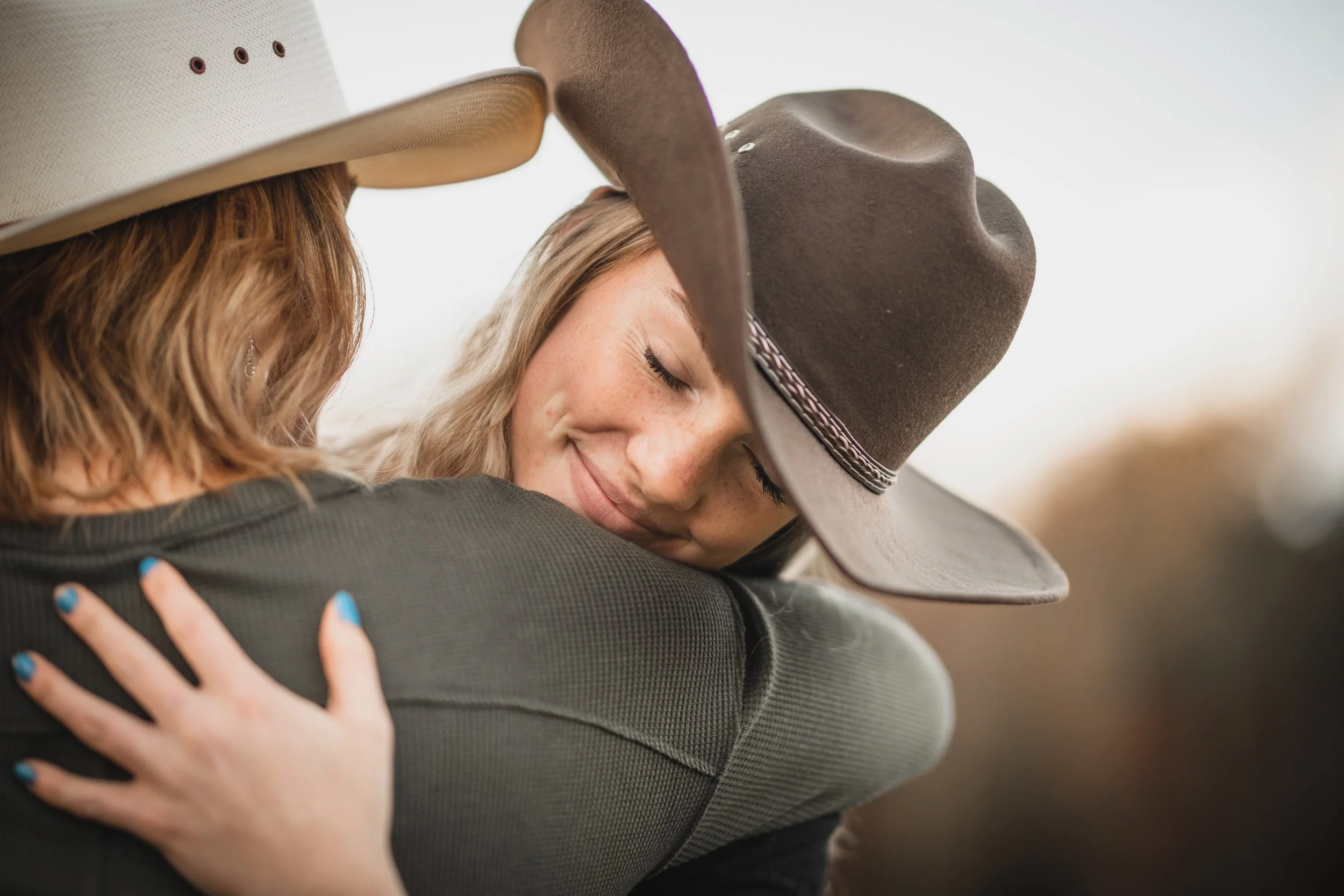 Two women wearing cowboy hats hugging outdoors, smiling with eyes closed.