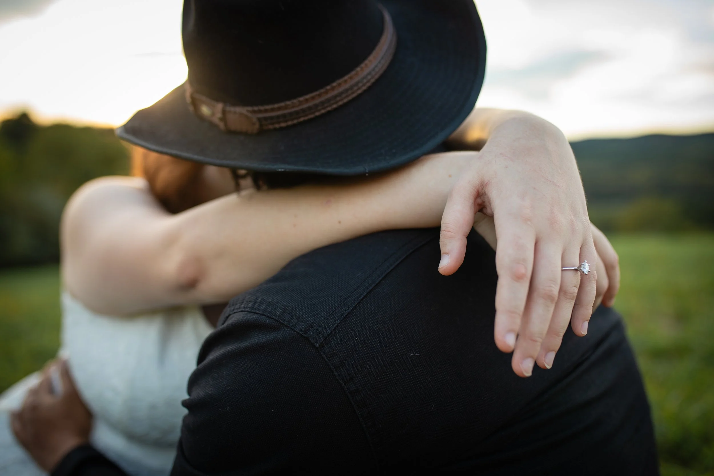 A woman and man embracing outdoors, the woman's arms around the man's neck, wearing a ring and a black hat, with a blurred natural landscape background.