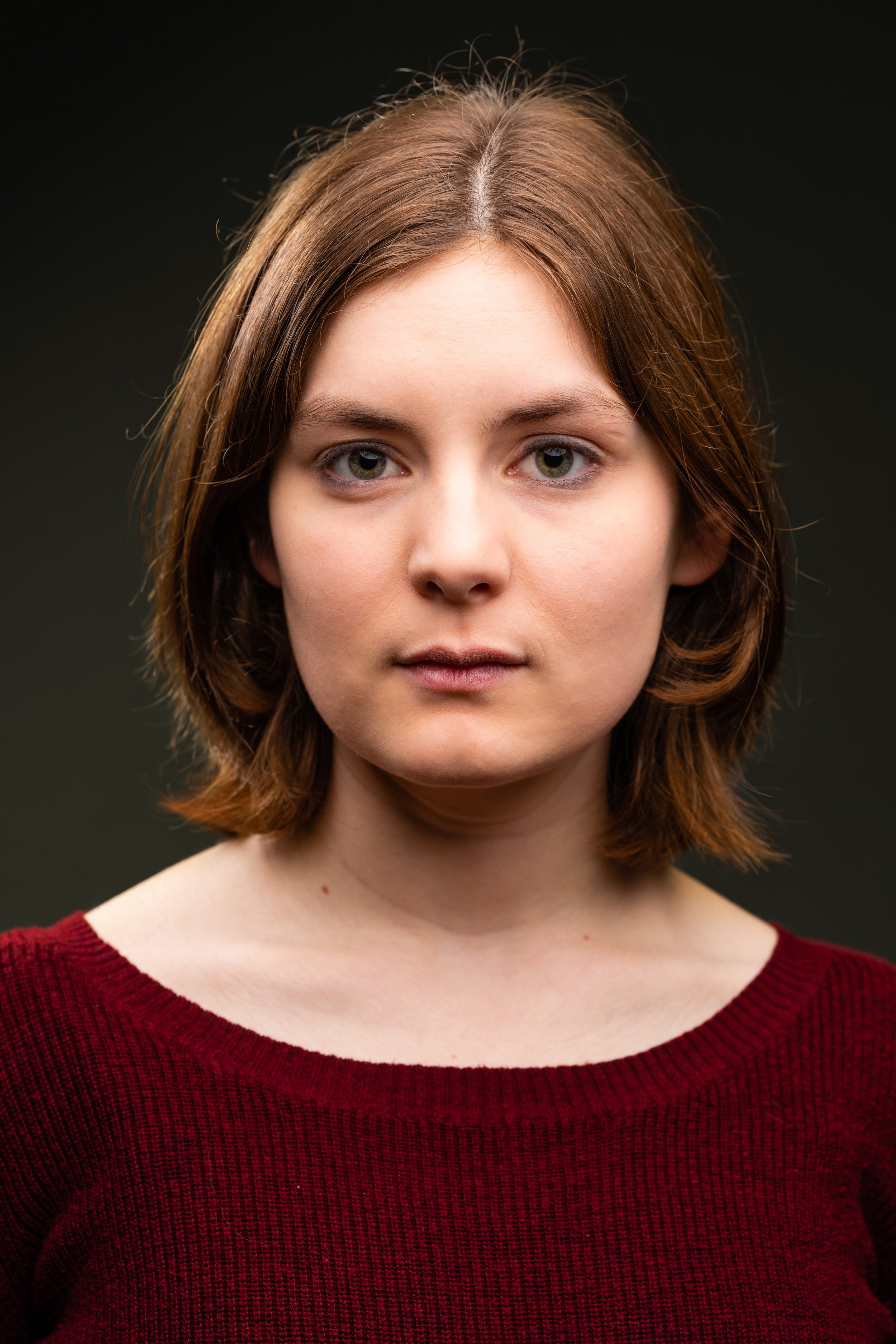A young woman with shoulder-length light brown hair, wearing a red sweater, and looking directly at the camera against a dark background.