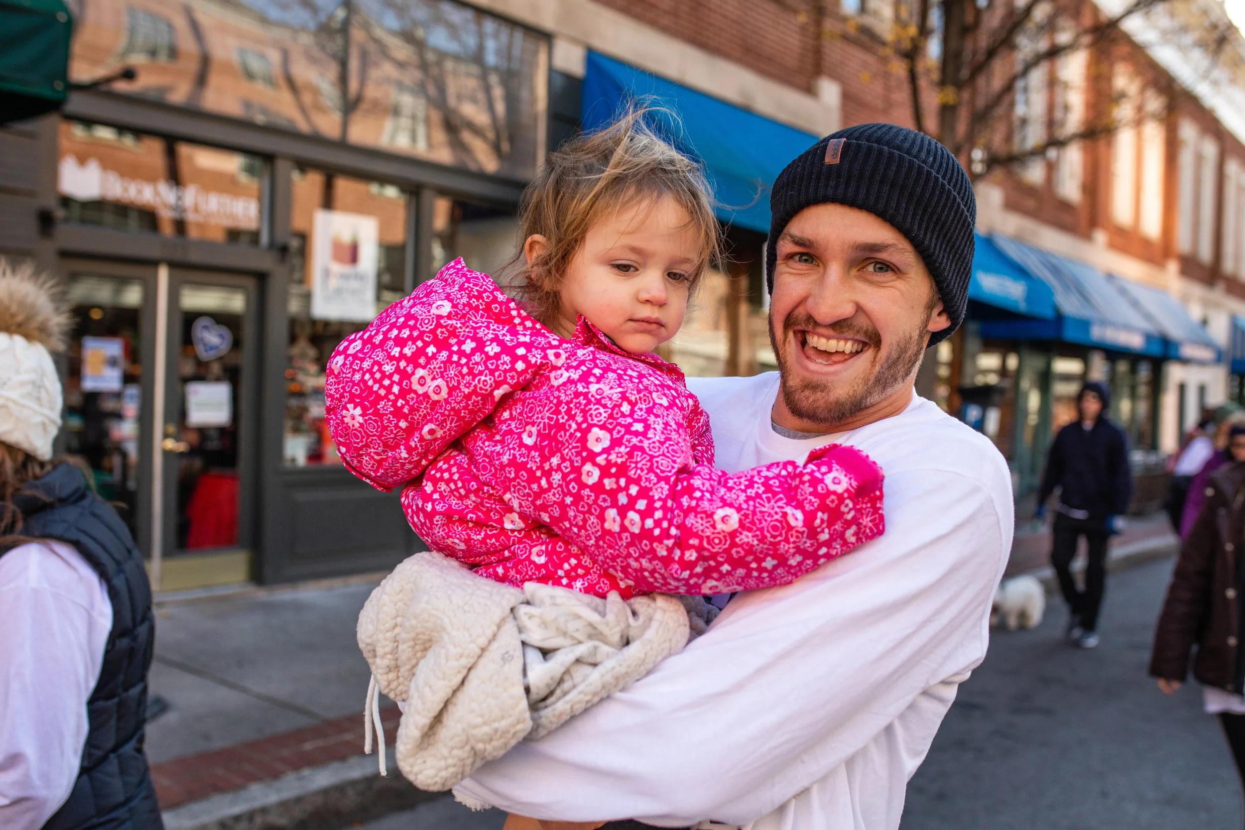 A man smiling and holding a young girl in a pink patterned jacket on a city street during daytime.