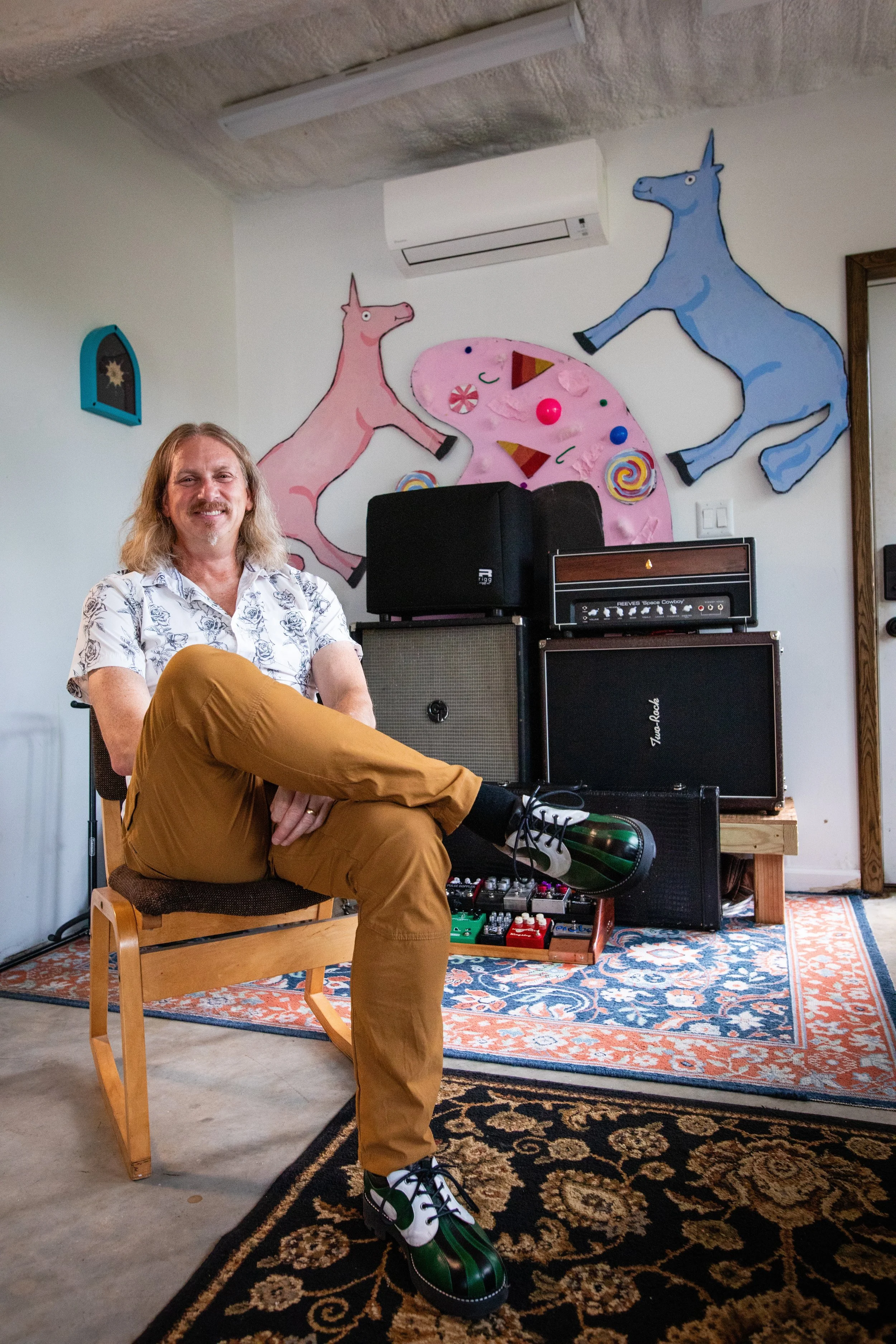 Man sitting in a music rehearsal space with musical equipment and colorful wall art of pink and blue unicorns.
