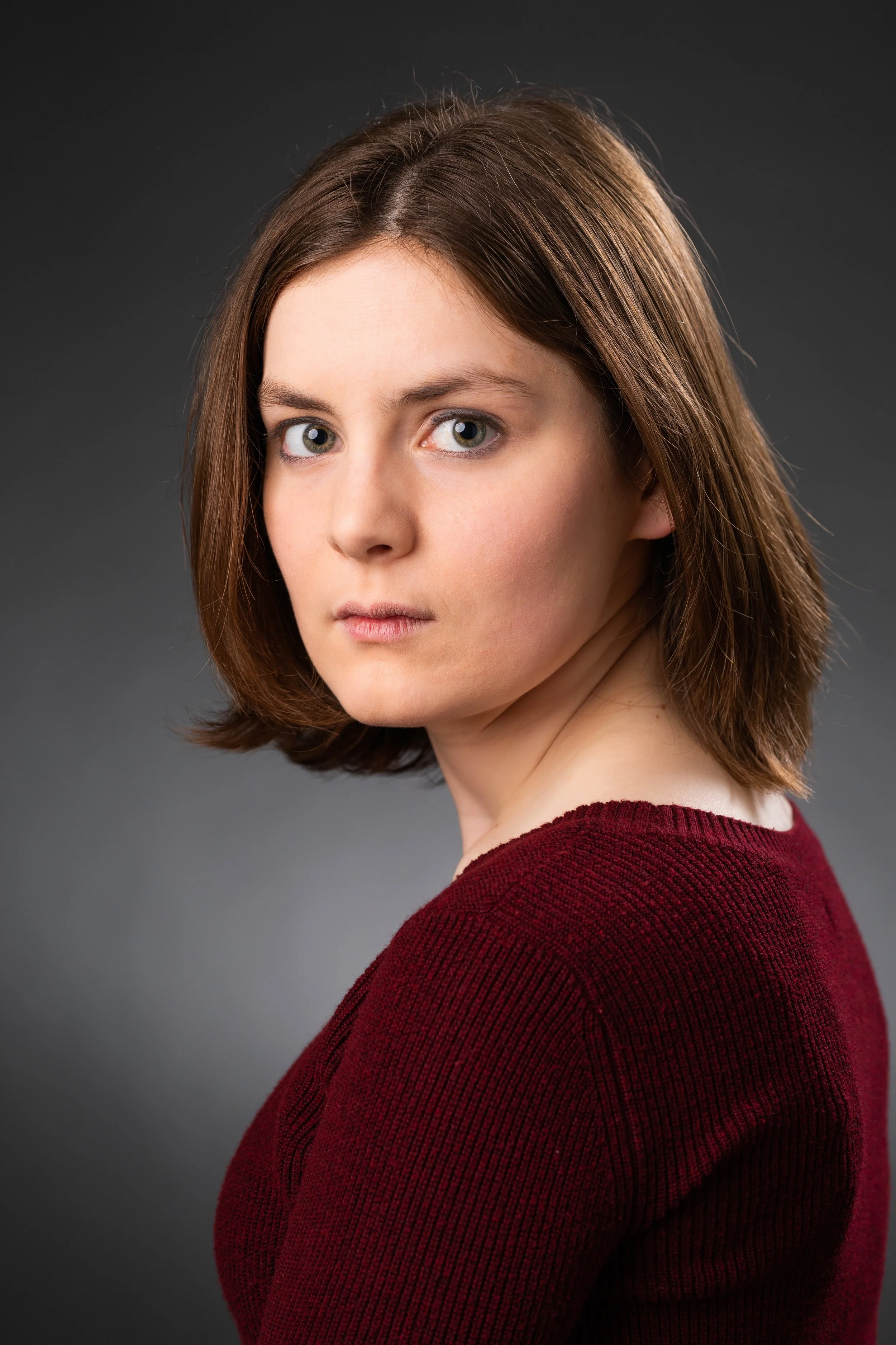 Close-up portrait of a young woman with shoulder-length brown hair, wearing a dark red sweater, against a gray background.
