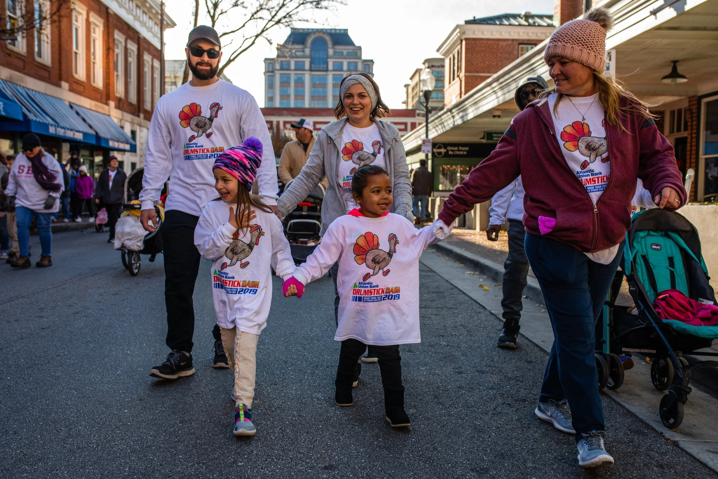 Group of people, including children and adults, walking and holding hands during a parade or event on a city street. Many are wearing white t-shirts with a turkey graphic and text, suggesting participation in an event like a turkey trot or fun run.