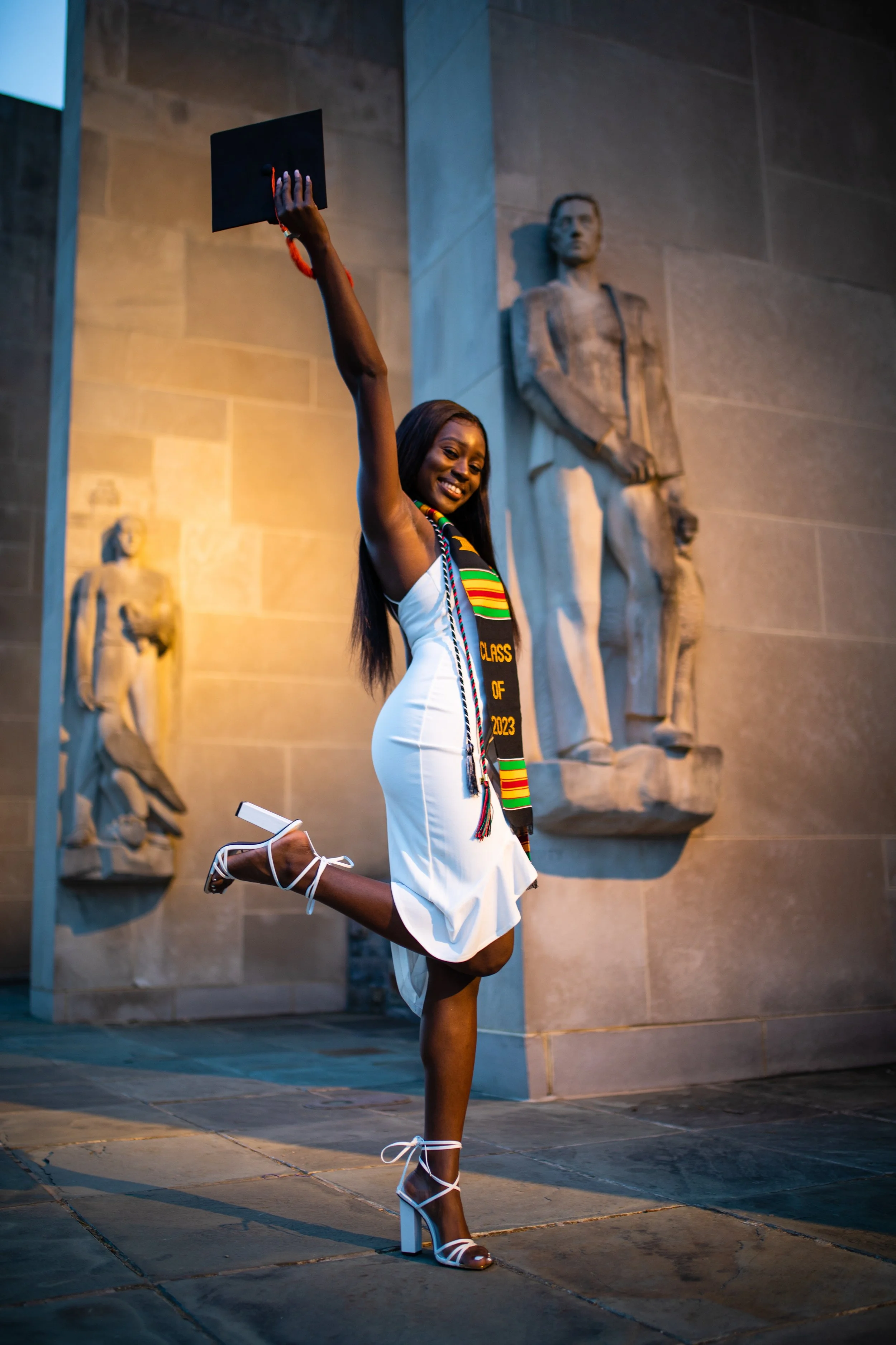 A young woman in a white dress and high heels celebrating graduation outside, holding her cap in the air while smiling, with statues on the wall behind her.