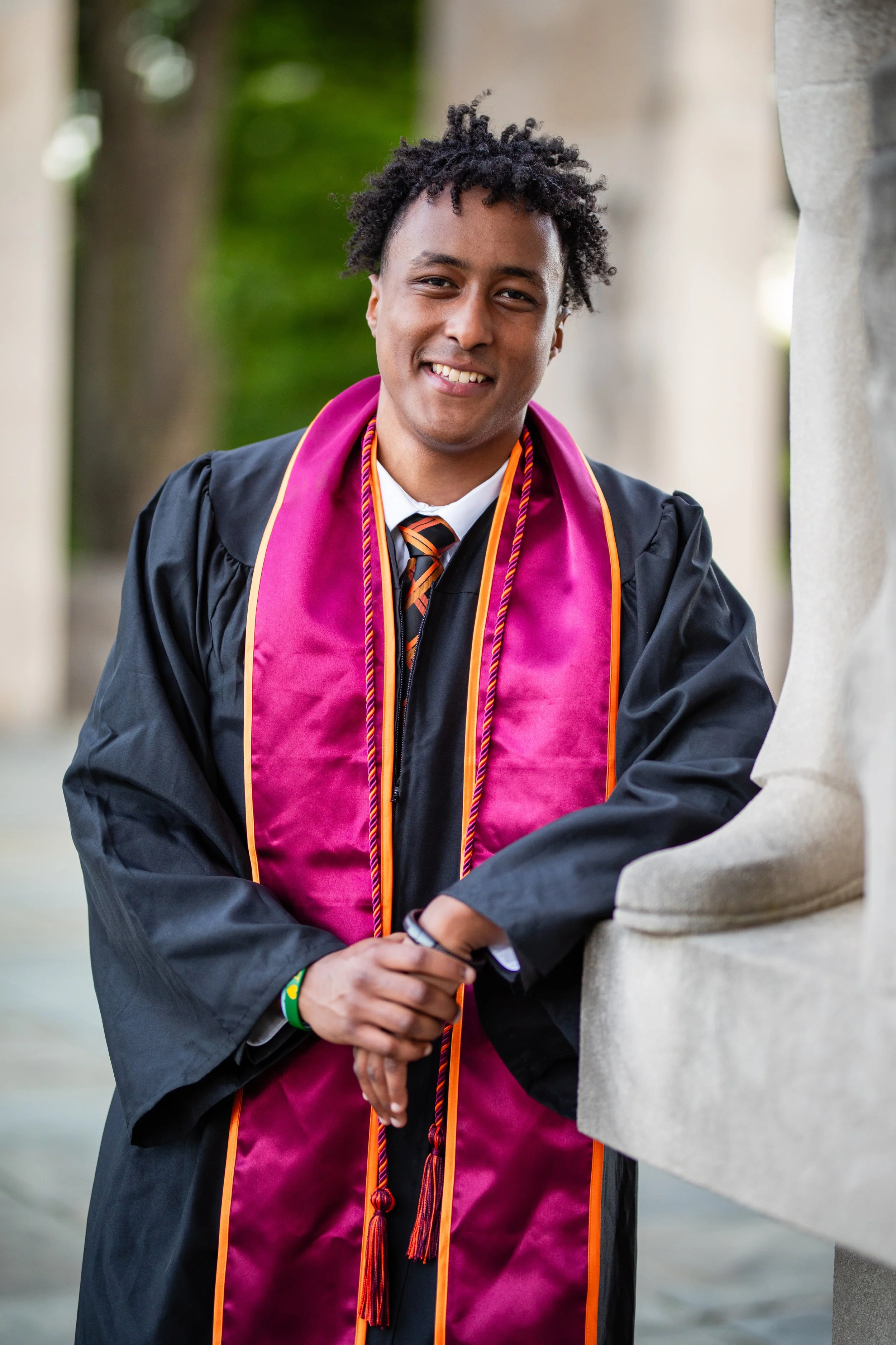 A young man in a graduation cap and gown with a pink stole and honor cords smiling outdoors.