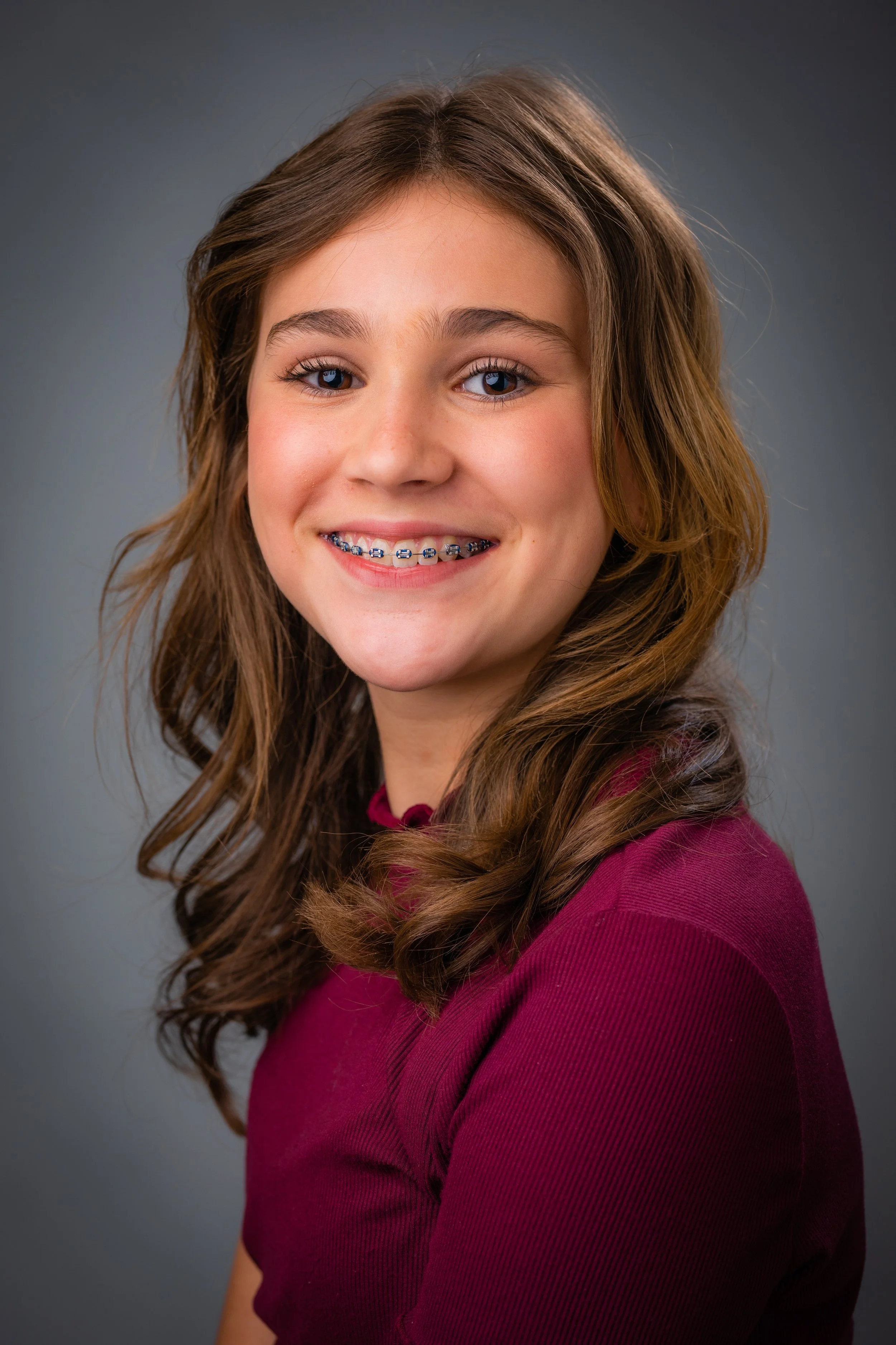 A young girl with long, curly brown hair, blue eyes, and braces on her teeth, wearing a maroon shirt, smiling at the camera against a gray background.