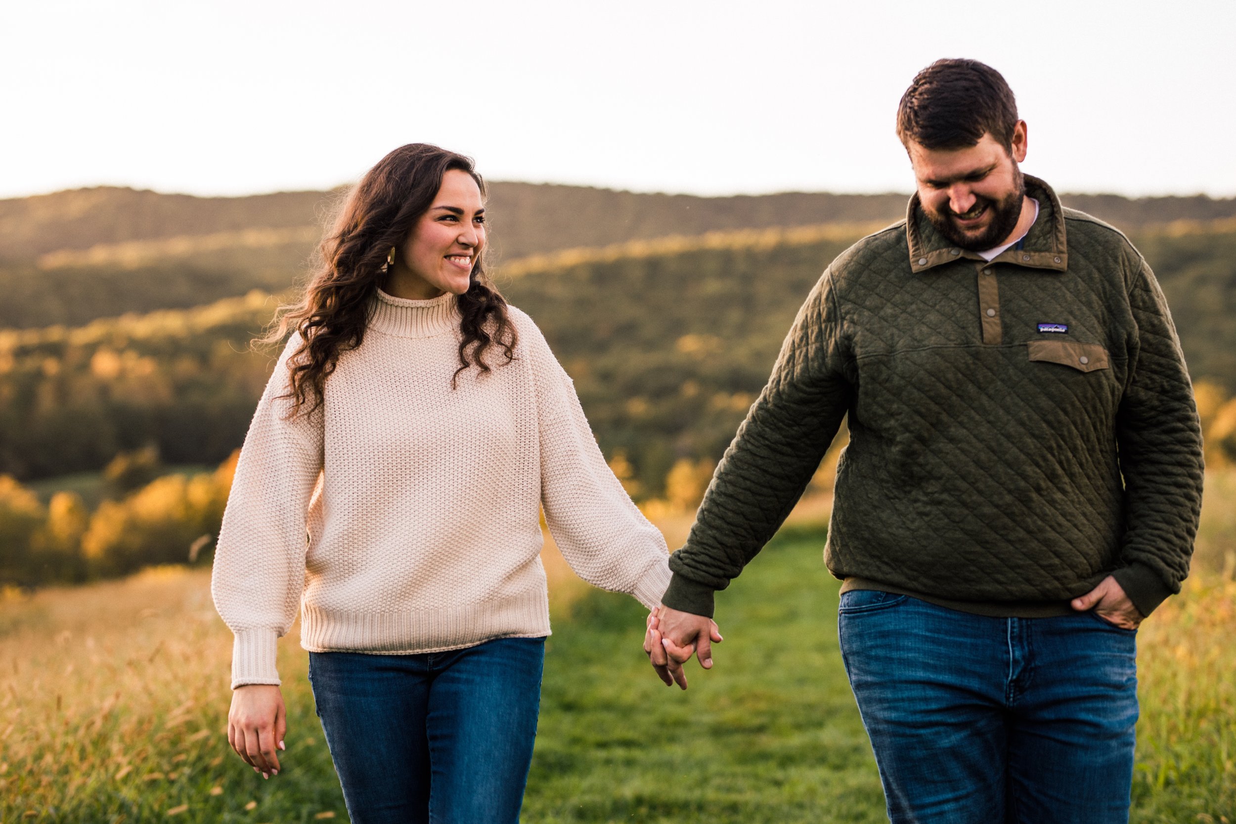 A smiling woman and man holding hands while walking outdoors in a grassy field with hills in the background during sunset.