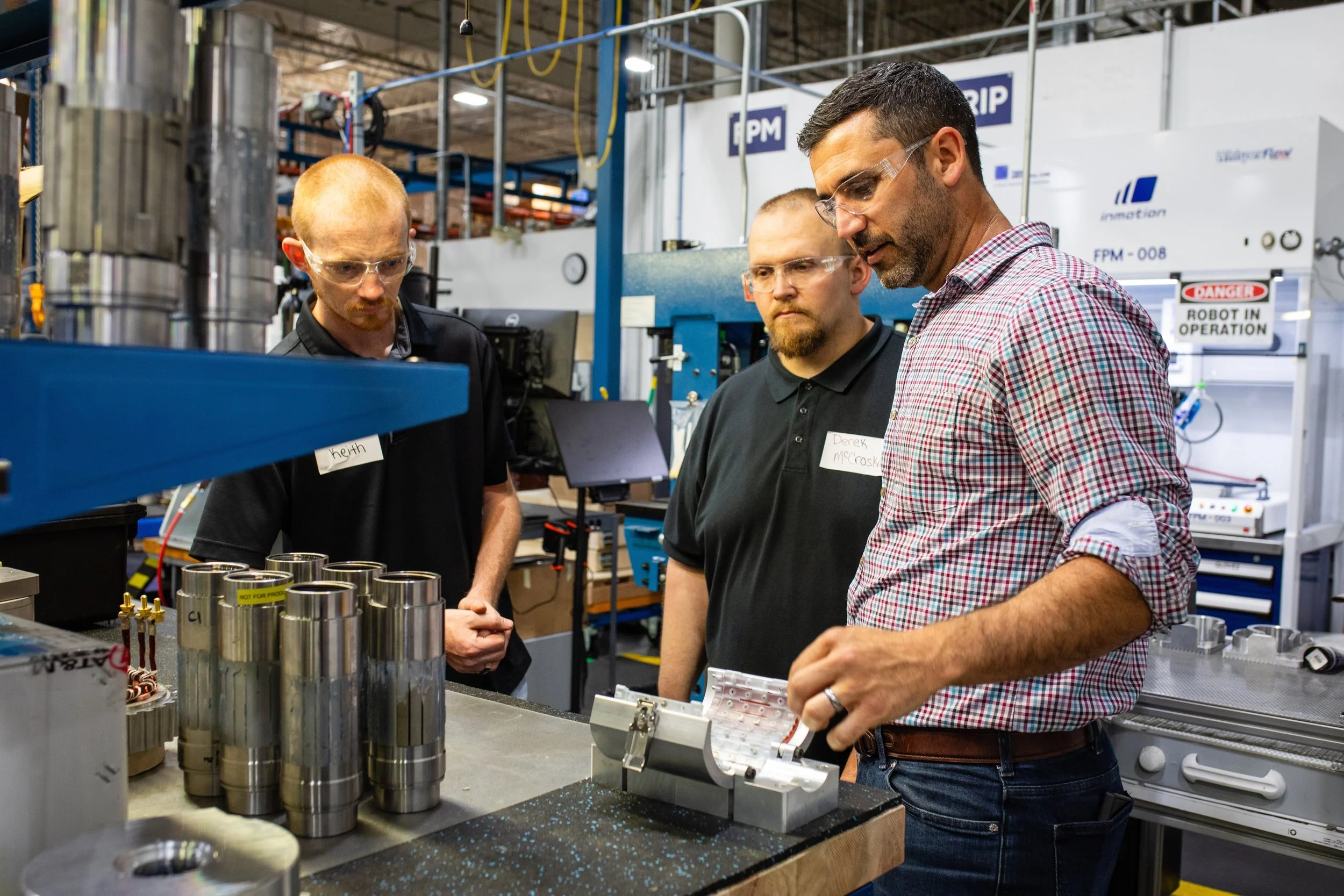 Three men working in a manufacturing or engineering lab, inspecting metal parts and machinery.