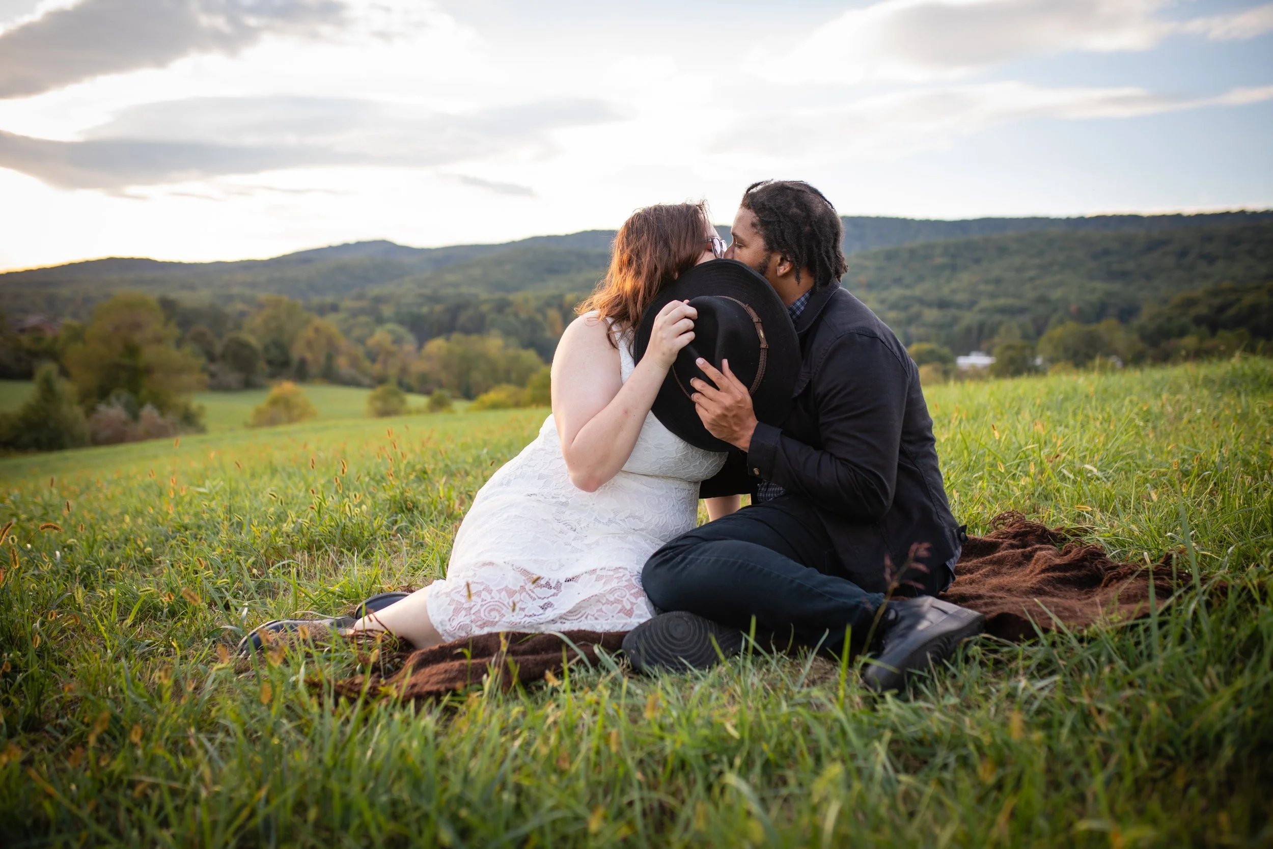 A couple sharing a kiss outdoors on a grassy field with hills and trees in the background.