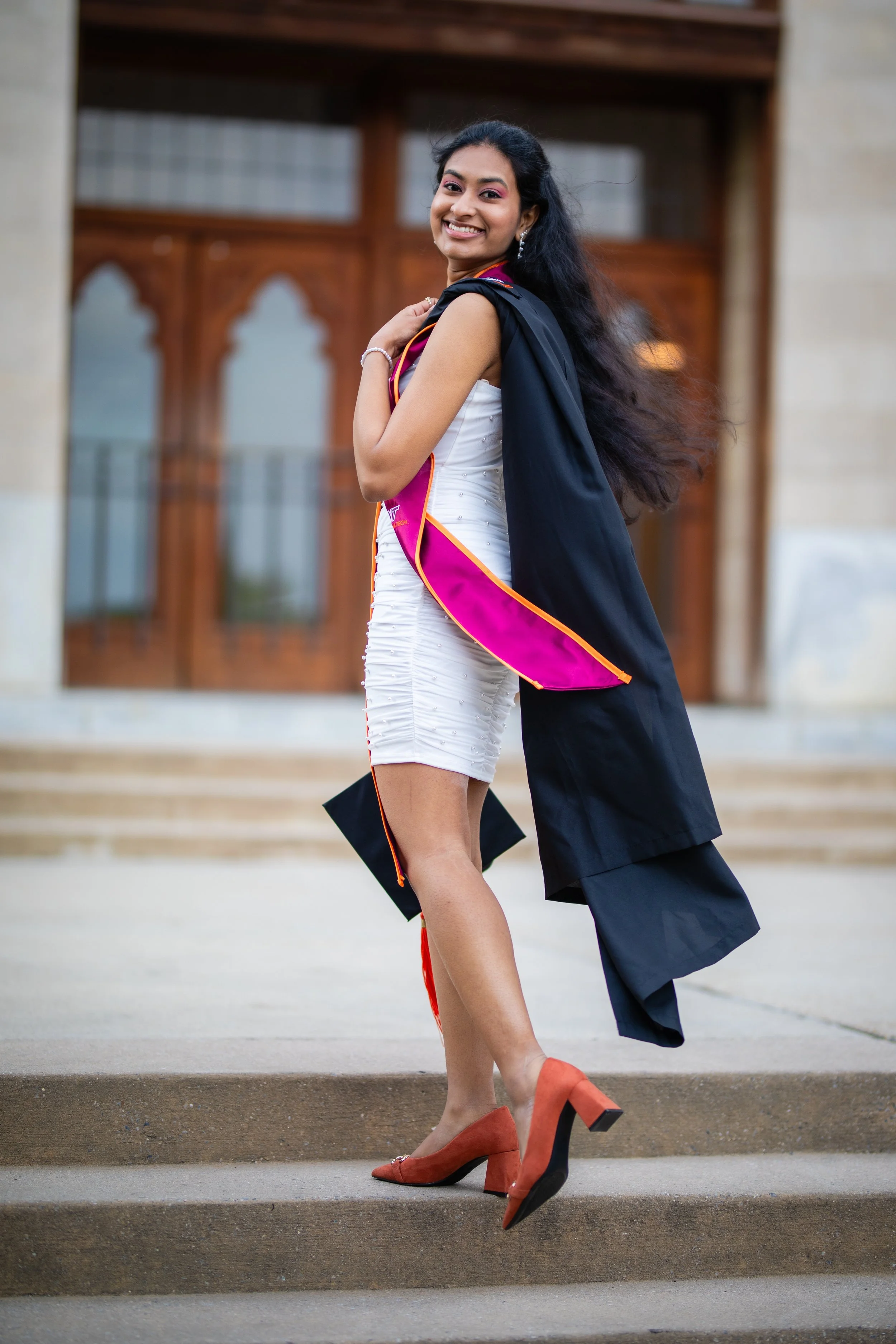 A young woman in a white dress and orange heels celebrating graduation, standing on steps in front of a building.