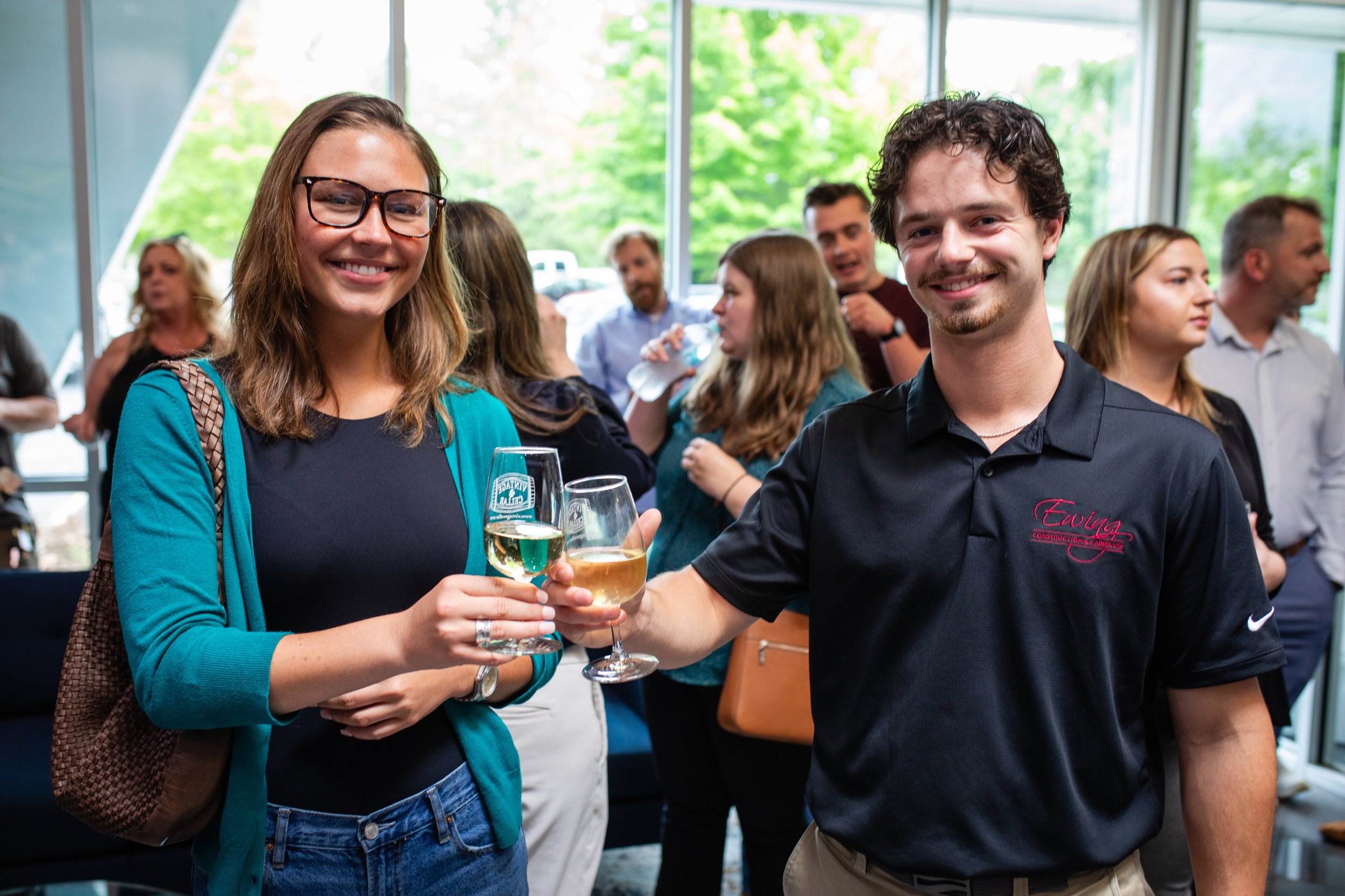 Two young adults smiling and toasting with wine glasses at a social gathering indoors with large windows and green trees outside.