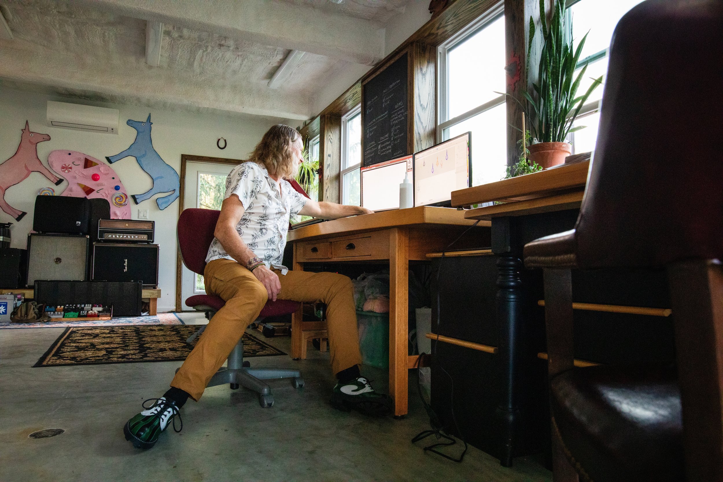 A person sitting at a wooden desk with two monitors, working or browsing online. The room has large windows, potted plants, and music equipment against the wall with colorful unicorn and donut artwork.