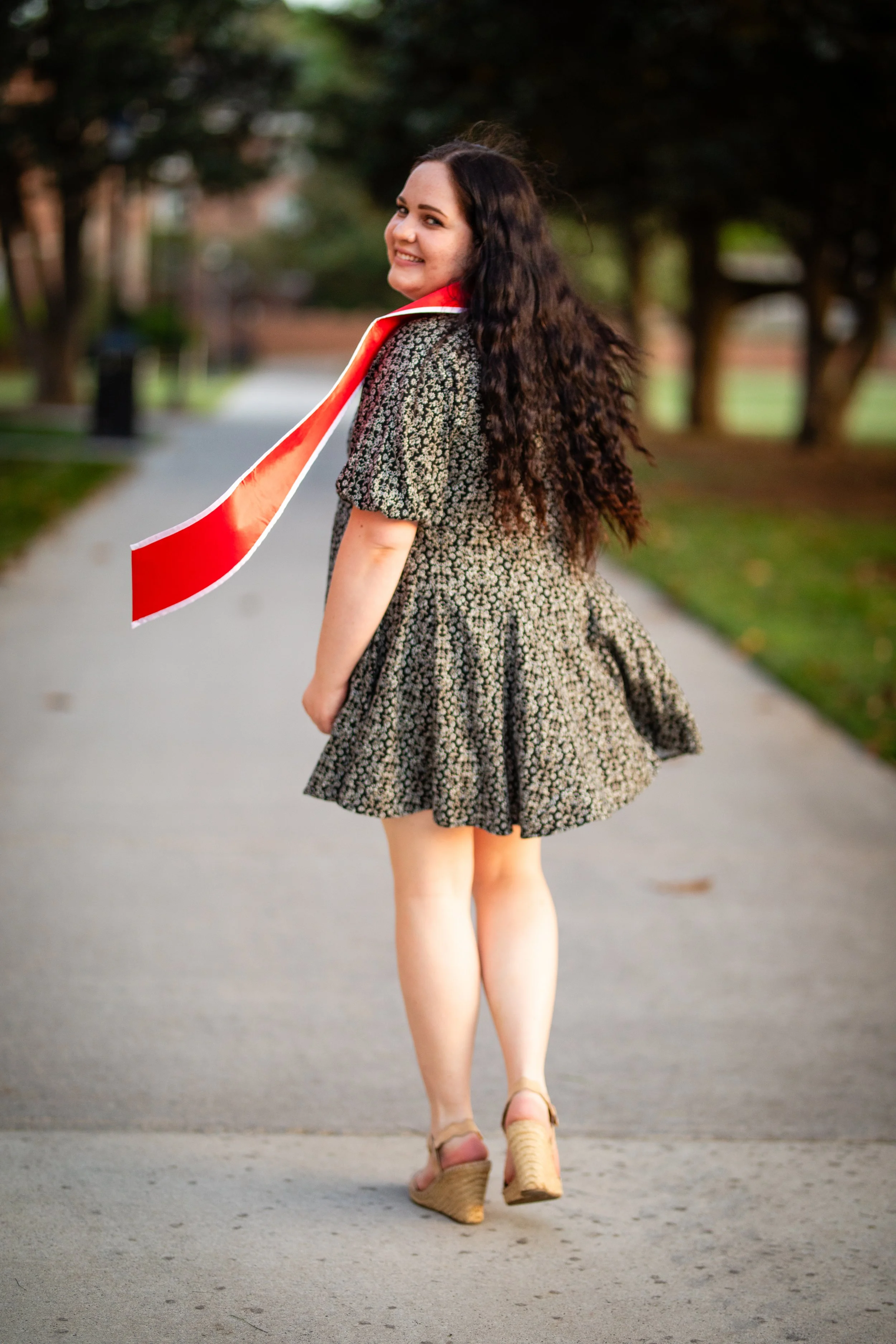 A young woman with long, dark, curly hair walking outdoors on a paved path, wearing a floral dress, a red and white scarf, and beige wedge sandals, smiling and looking over her shoulder.