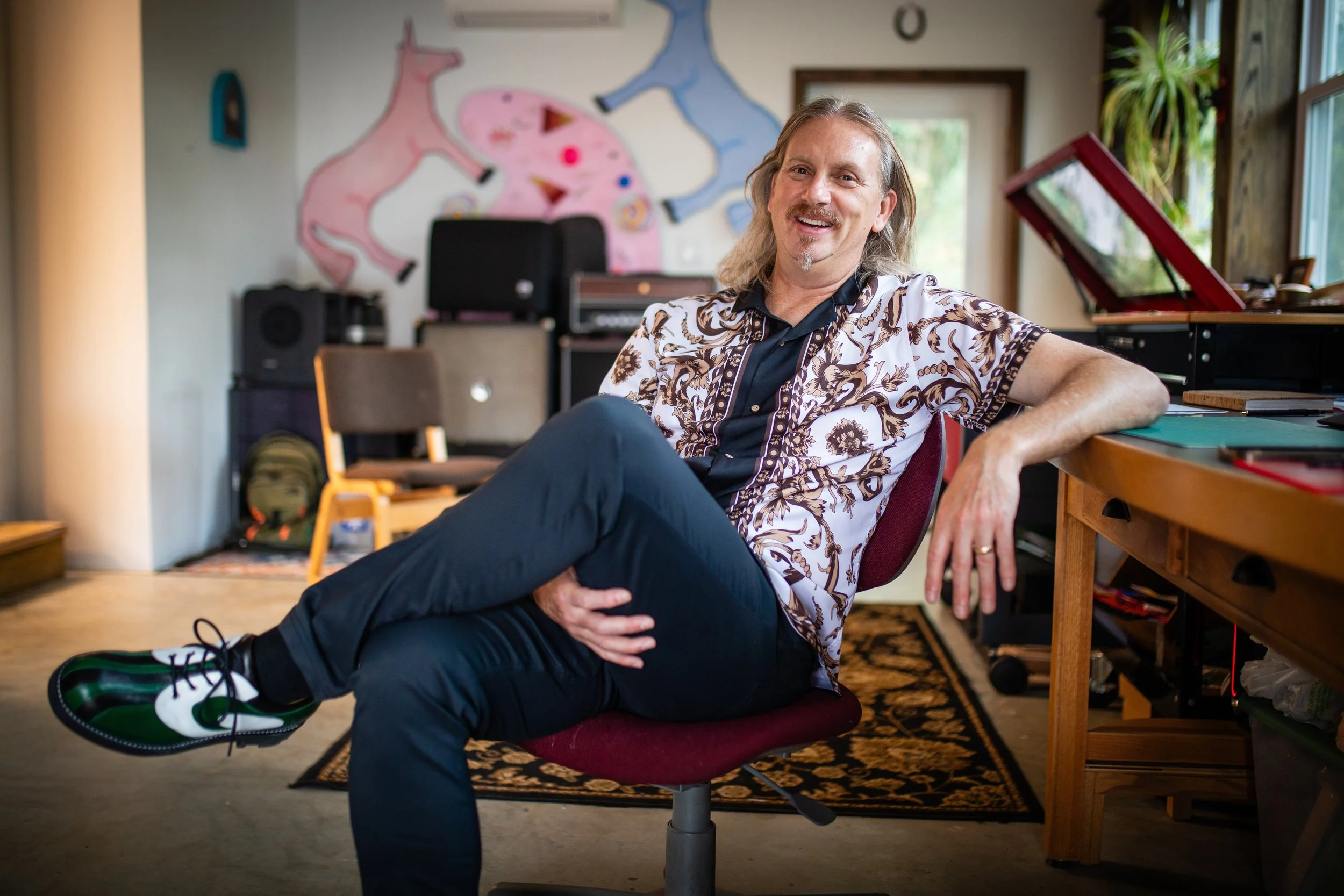 A man with long gray hair and a mustache, smiling, seated on a red office chair in a room with musical equipment and artwork on the wall.