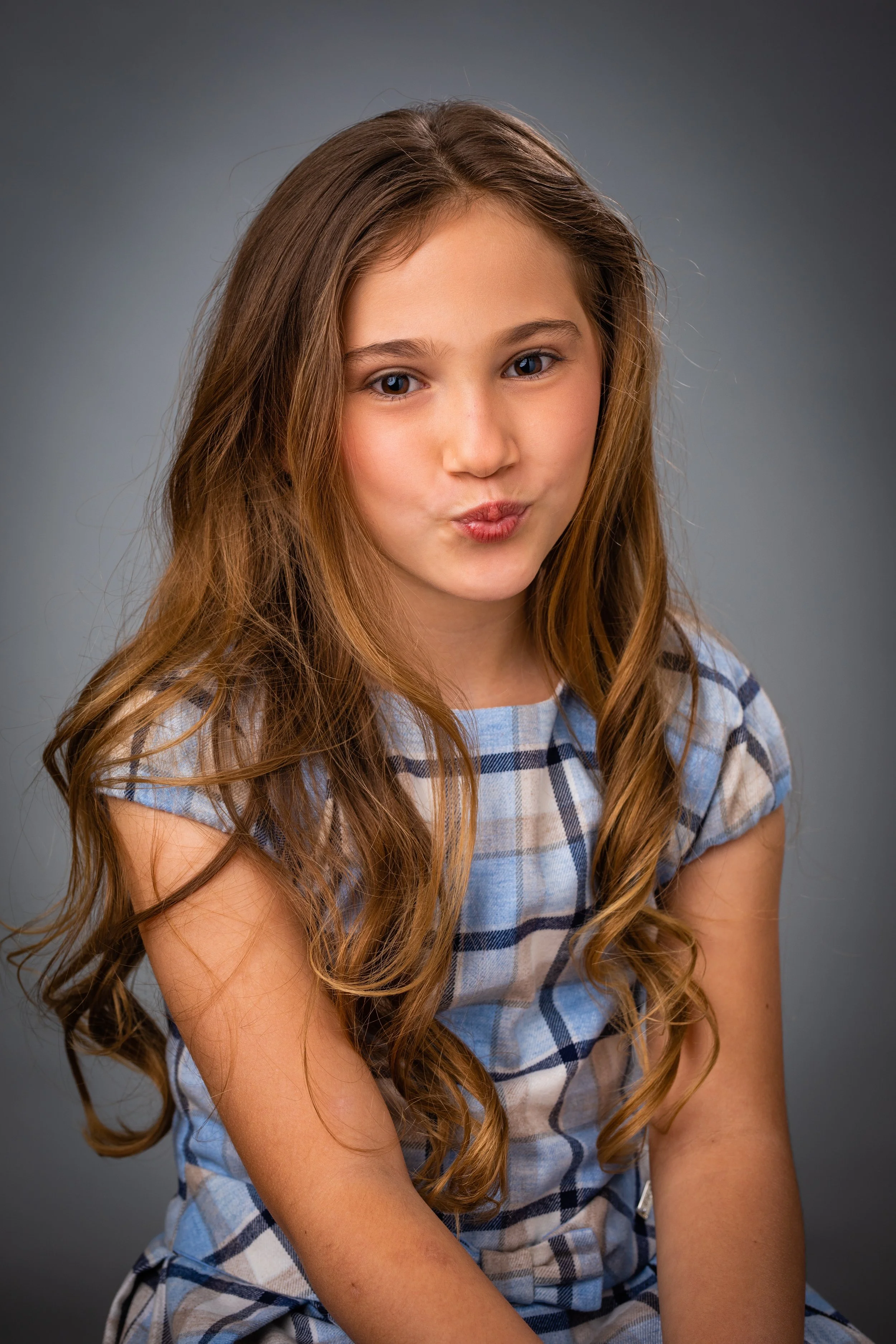 A young girl with long, wavy brown hair wearing a blue and gray plaid dress, looking at the camera with a playful expression against a plain gray background.