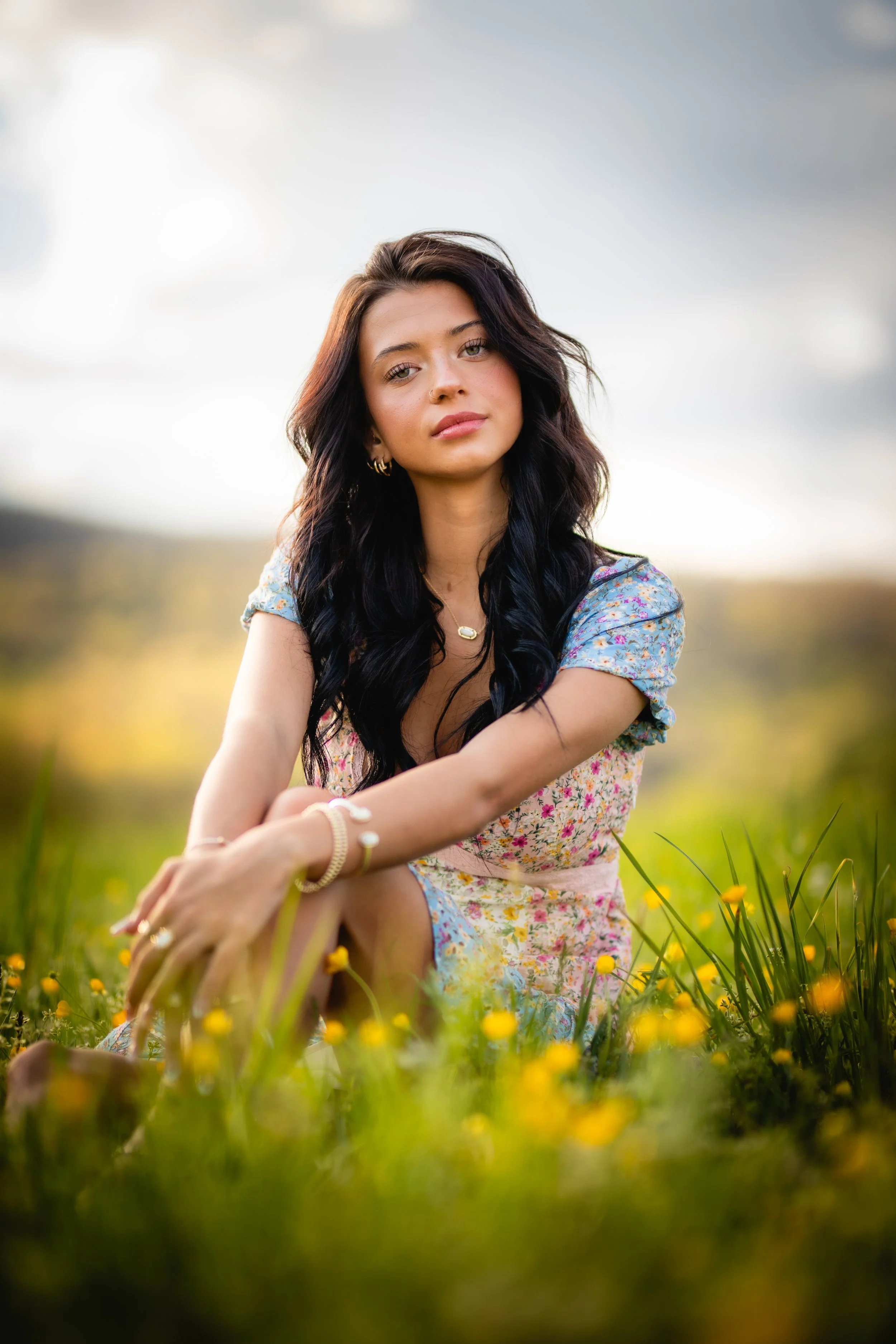 A young woman with dark wavy hair and light skin sitting in a field of yellow flowers, wearing a floral dress and jewelry, looking at the camera with a calm expression.