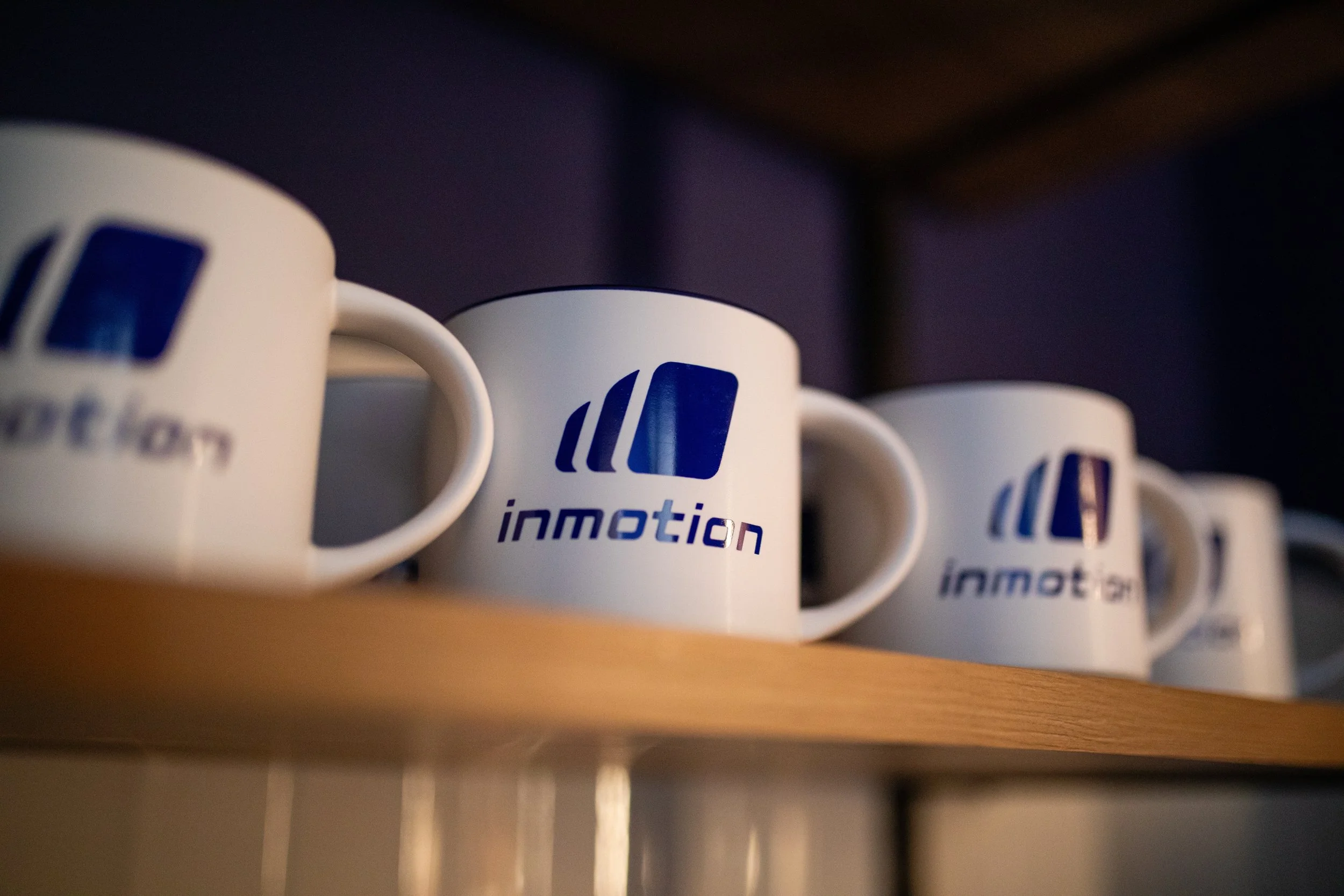 White coffee mugs with blue logo and text 'inmotion' on a wooden shelf.