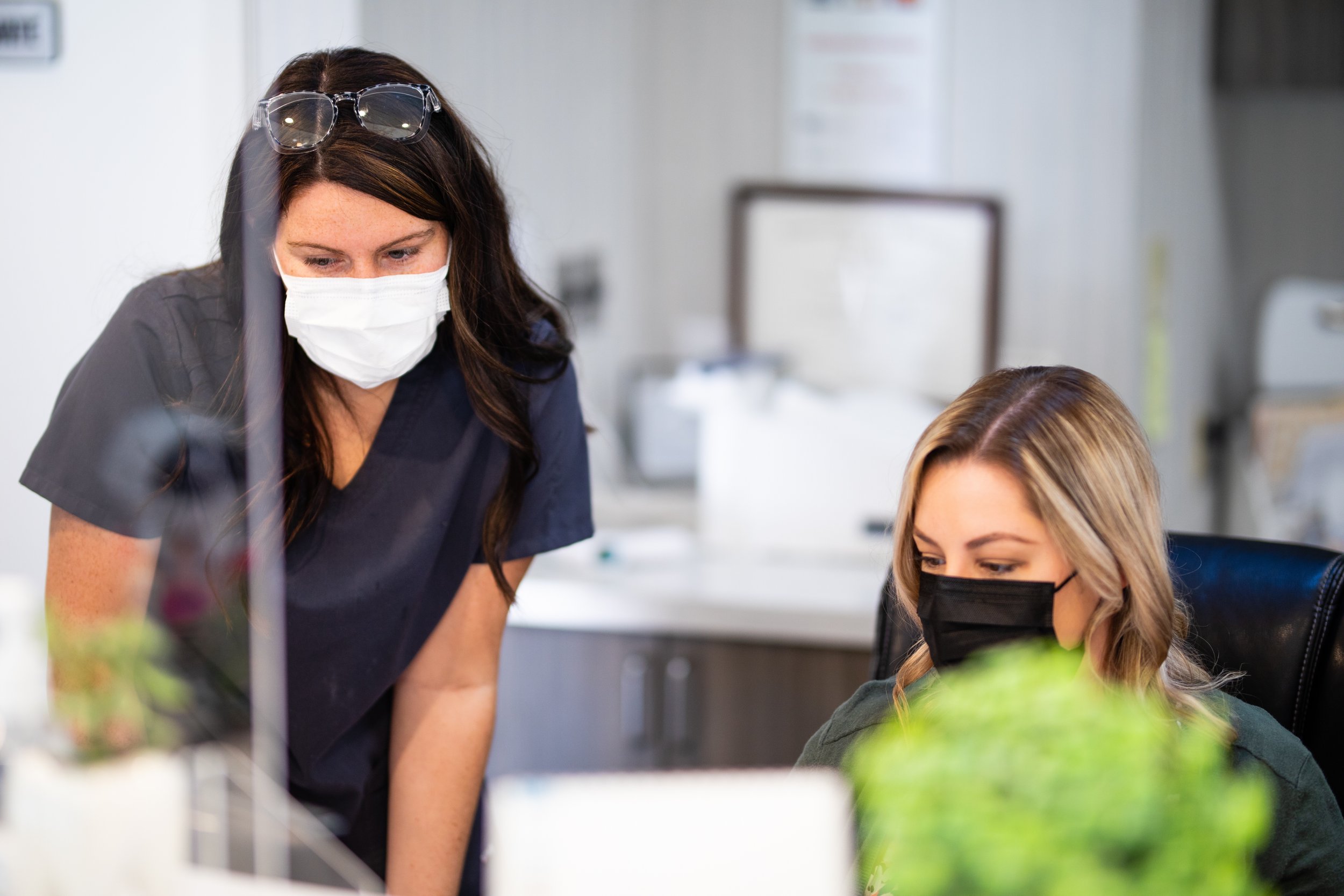 Two women in a medical or laboratory setting wearing face masks, one standing and one sitting, with blurred laboratory equipment in the background.