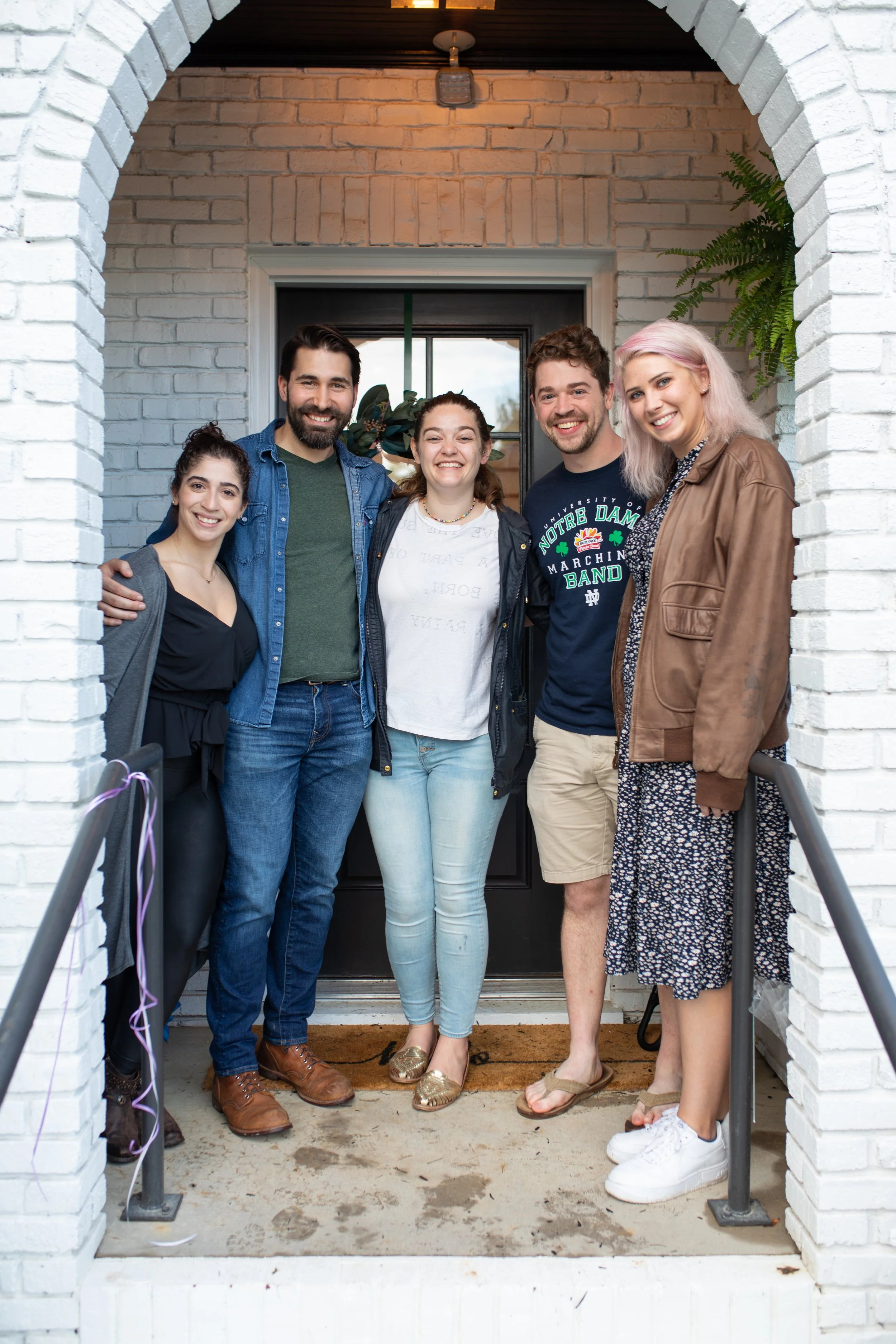 Group of five friends standing on porch, smiling at camera, in front of house door, brick wall, some with arms around each other, casual clothing, celebration setup.