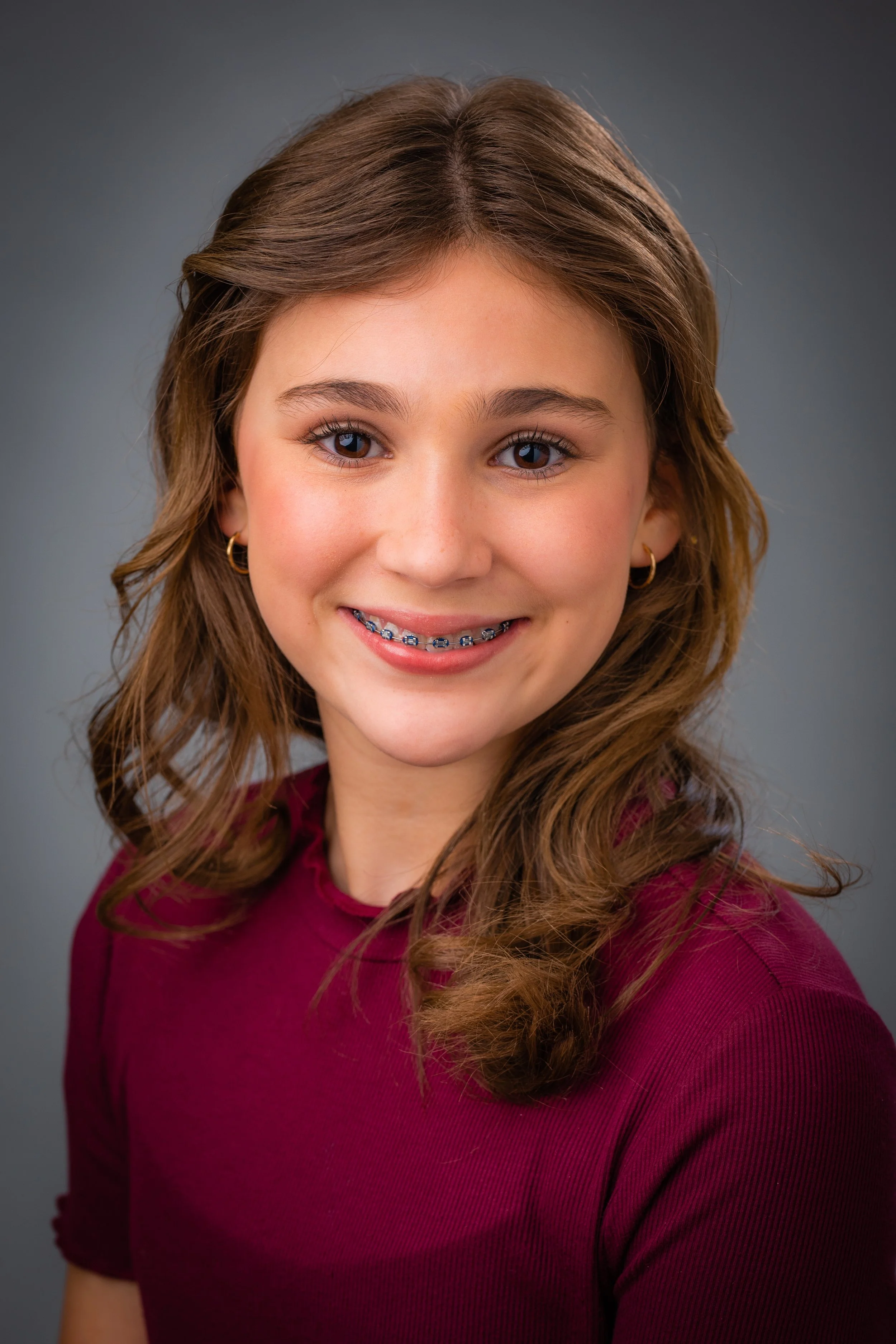 A young girl with wavy brown hair, braces, and blue eyes, wearing a maroon top and small hoop earrings, smiling against a gray background.