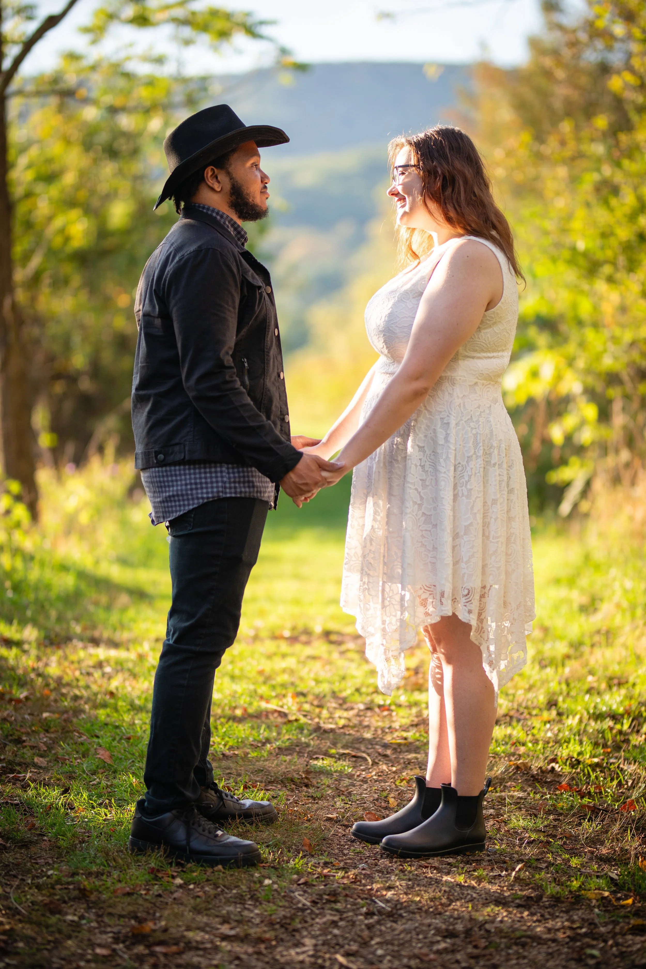 A couple holding hands and gazing at each other outdoors on a sunny day, with trees and mountains in the background.