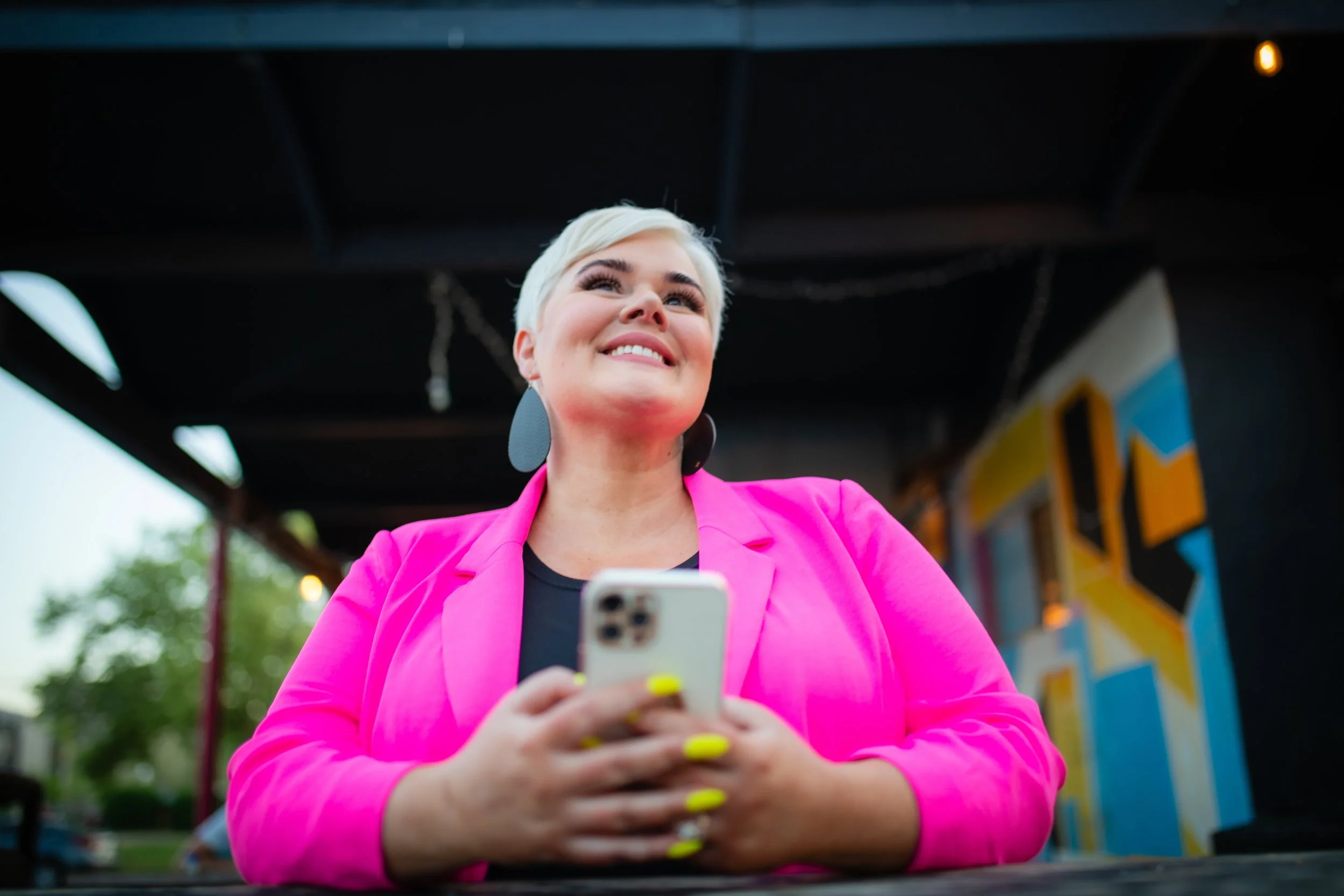 Woman with short blonde hair wearing a bright pink blazer and black top, smiling and looking up while holding a smartphone with yellow-painted nails, standing outside near colorful graffiti art.