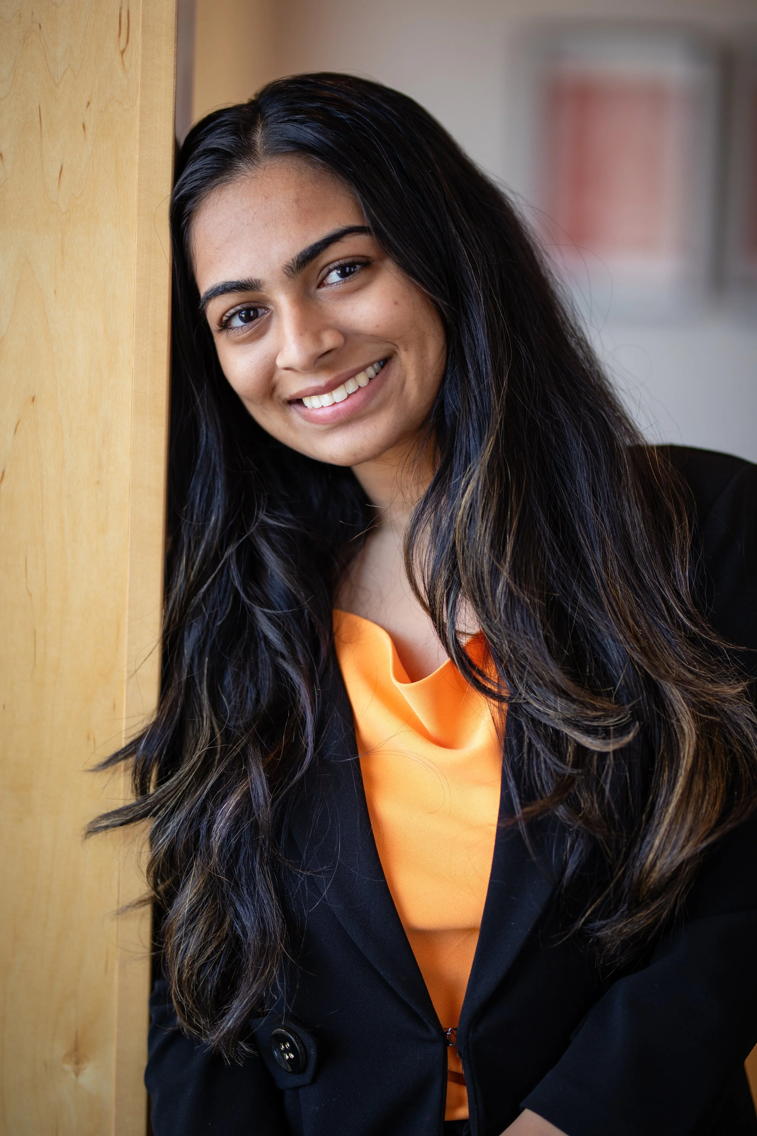 A woman with long dark hair smiling and leaning against a wooden surface, wearing a black blazer over an orange top.