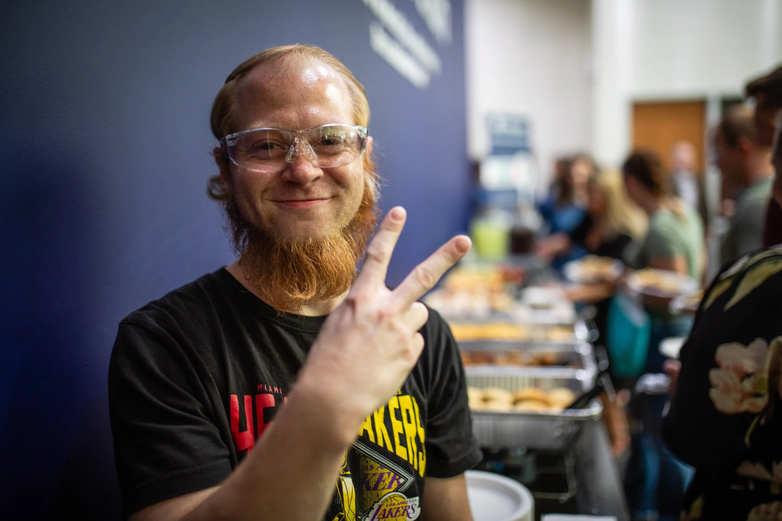 A smiling man with glasses and a red beard making a peace sign with his right hand at a buffet line, with other people and food in the background.