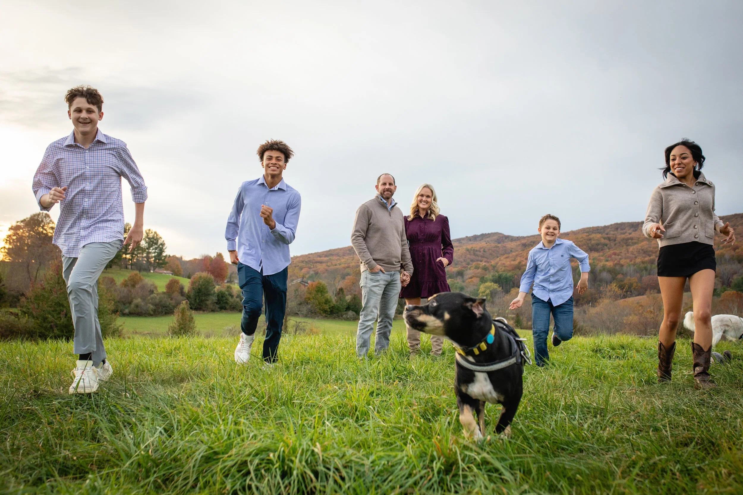 A family of six and their dog running and playing on a grassy field with a scenic autumn landscape and hills in the background.