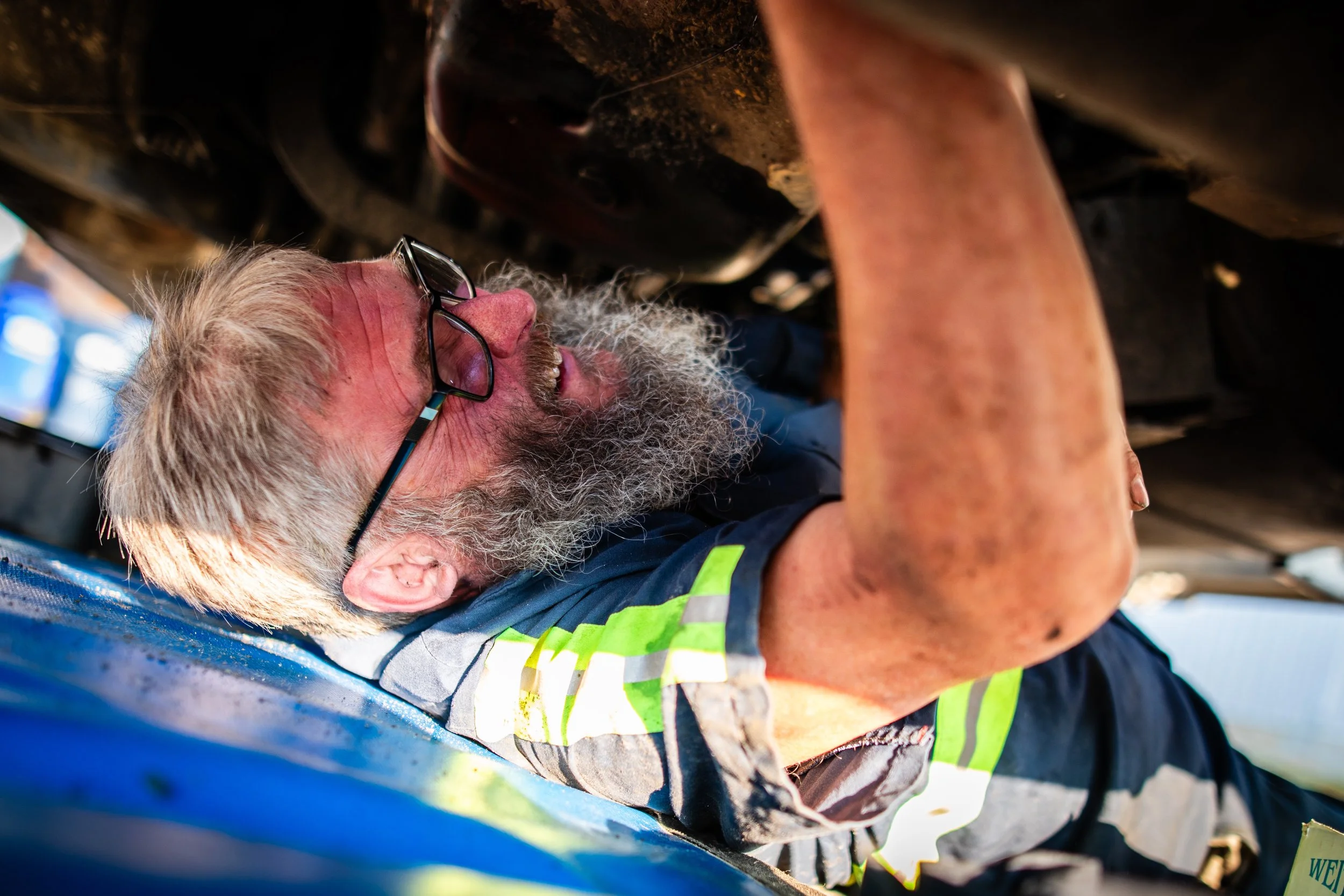 A man with gray hair, a beard, and glasses working underneath a vehicle in a workshop or garage.