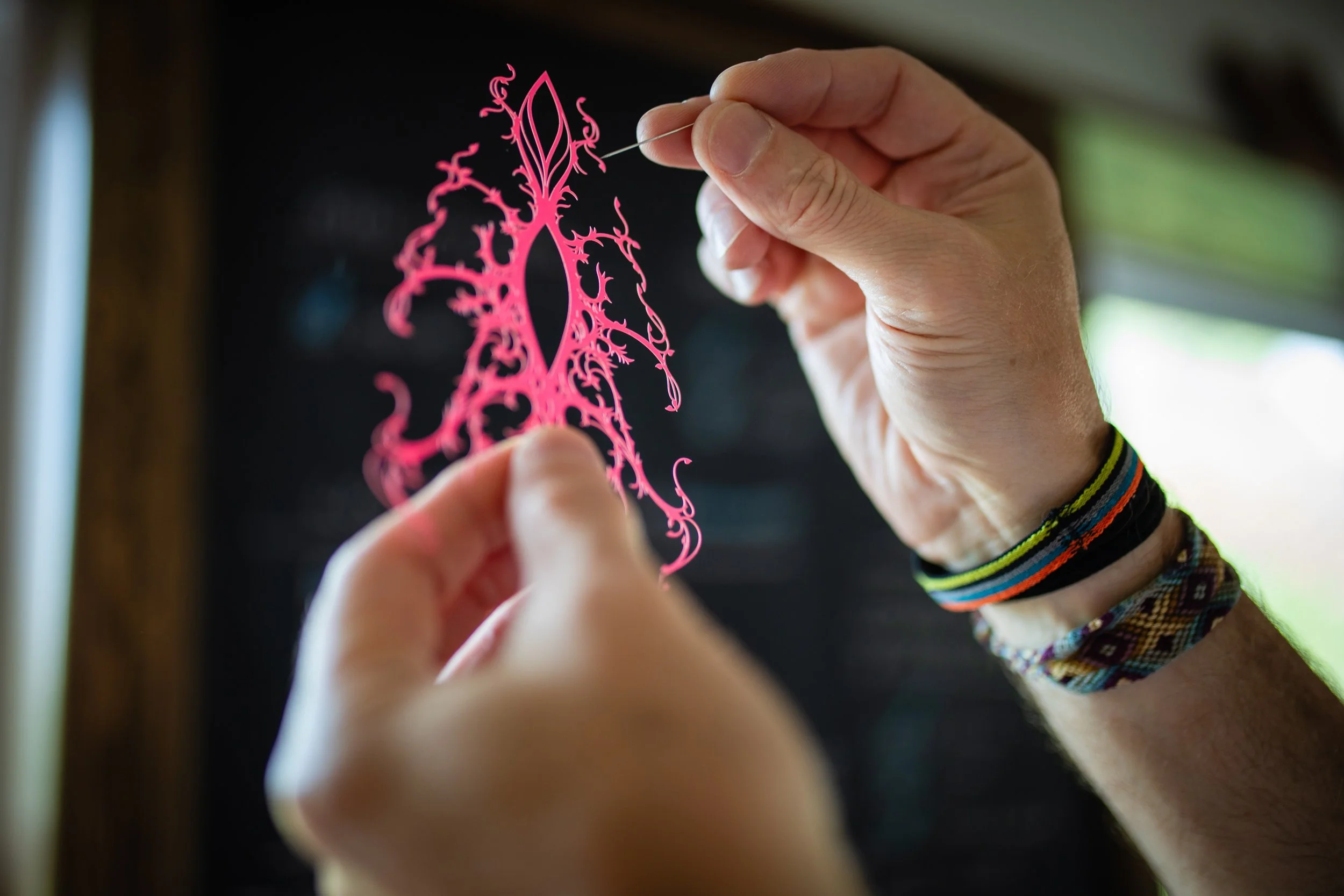 Person holding pink paper cutout with elaborate, intricate design in front of a dark background with a window and blurry outdoor scenery.