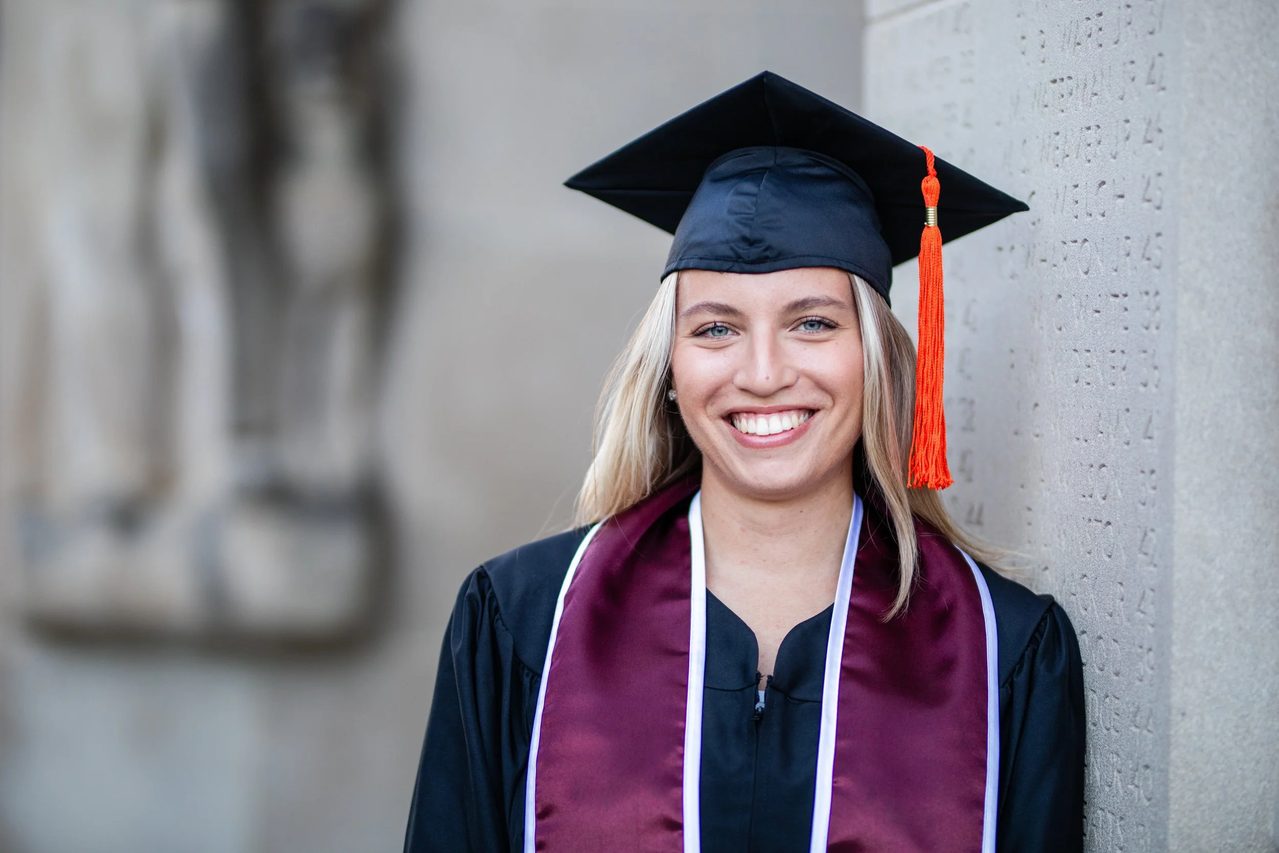 A young woman wearing a graduation cap and gown, smiling with joy, standing against a stone wall.